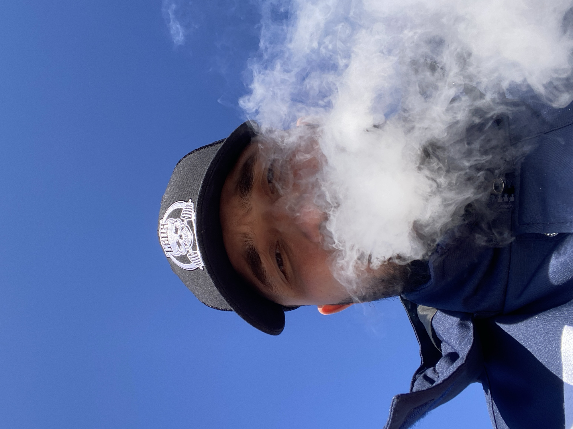 The image is a low-angle shot of a person looking downwards, set against a clear, bright blue sky. The person, appearing to be male, is largely obscured by a dense, white cloud of vapor or smoke emanating from their mouth and nose.
The subject is wearing a dark blue jacket or shirt and a dark baseball cap. On the front of the cap, there is a prominent white embroidered logo featuring a skull surrounded by what appears to be barbells or weights. Partially visible text above the skull seems to include the letter "H" and potentially spells out "CRUSH".
The vapor obscures the lower half of the person's face, making it difficult to discern their expression, though their eyes are visible above the smoke. The bright blue sky suggests an outdoor setting during the daytime with clear weather. The overall activity depicted is the exhaling of a large cloud of vapor, likely from vaping. The image provides no specific location cues, but the context indicates it was taken in Luzhitsy, Russia.