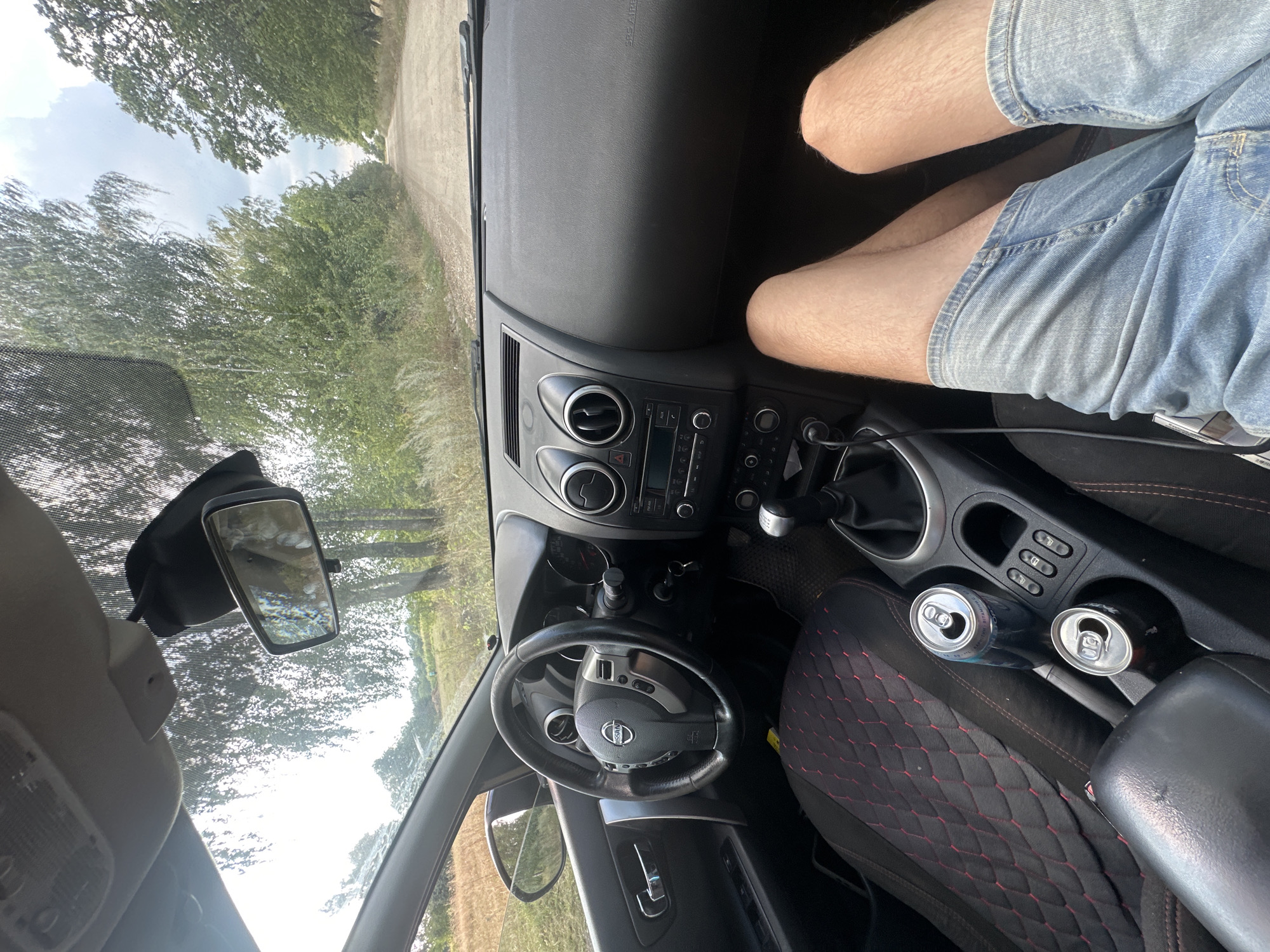 The image depicts the interior of a car looking out onto a dirt road lined with trees and fields. The perspective is from the driver's seat, with the steering wheel, dashboard, and gear shift visible. The passenger's legs, clad in denim shorts, are prominent in the foreground. Two open beverage cans are situated in the cup holders between the front seats. The car appears to be stationary, possibly on a rural road outside of Tynna, Ukraine, given the unpaved road and natural surroundings. The sky is overcast, suggesting potentially unfavorable weather.