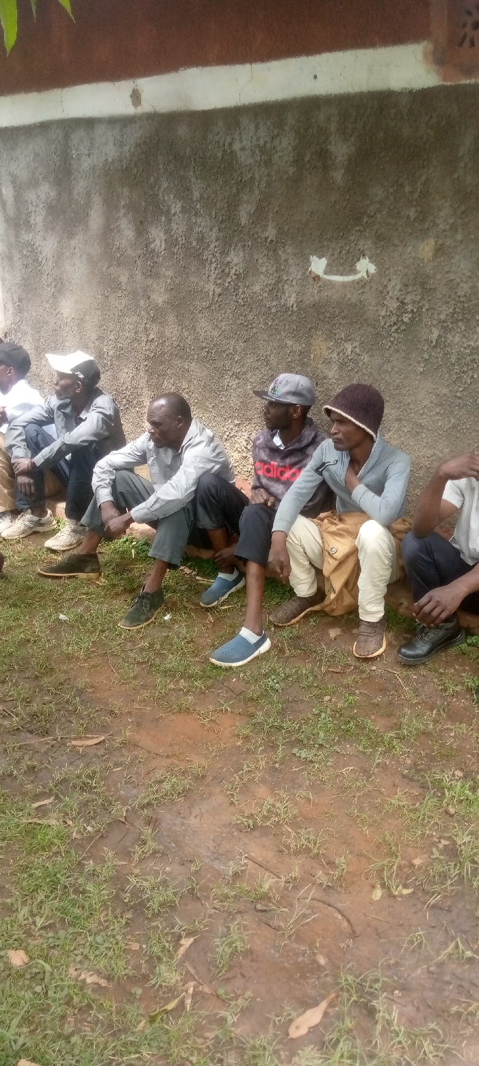 The image shows a group of five men sitting in a line on a dirt path in front of a concrete wall in Kivaywa, Kenya (latitude: 0.56, longitude: 34.806). The men are all wearing casual clothing and footwear. They are all looking in different directions. There is a small amount of green grass growing on the path. The concrete wall is cracked and weathered. There appears to be a white arrow painted on the wall.
