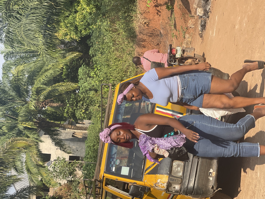 Two women rest on a yellow vehicle, possibly a taxi or van, in a tropical setting. One woman, wearing a light blue top and jean shorts, props herself up while the other, in a black tank top and jeans, lies seemingly asleep with a purple floral top beside her head. Behind them, a man sits on a motorbike, partially obscured. Lush green palm trees and foliage surround the scene, with a glimpse of a building in the background. The ground appears reddish brown.