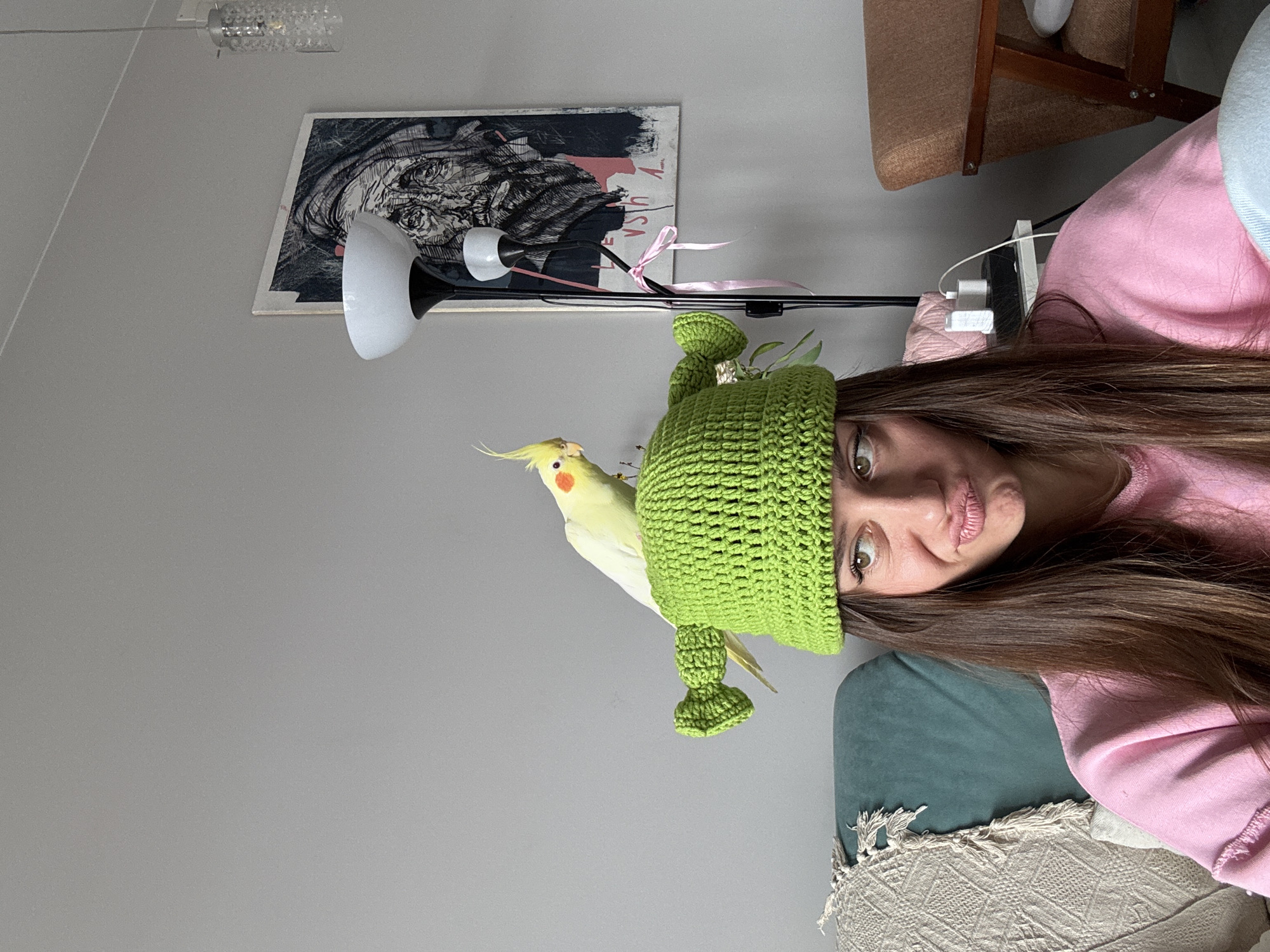 A woman is posing for a selfie with a cockatiel perched on a bright green, knitted hat on her head. The woman is wearing a pink long-sleeved shirt. The bird is predominantly white with a yellow head and orange cheek patches. In the background, there is a grey wall, a framed piece of art, a white lamp, and a brown chair. A USB charging block is also visible, as well as part of a throw blanket. The specific location is not indicated.