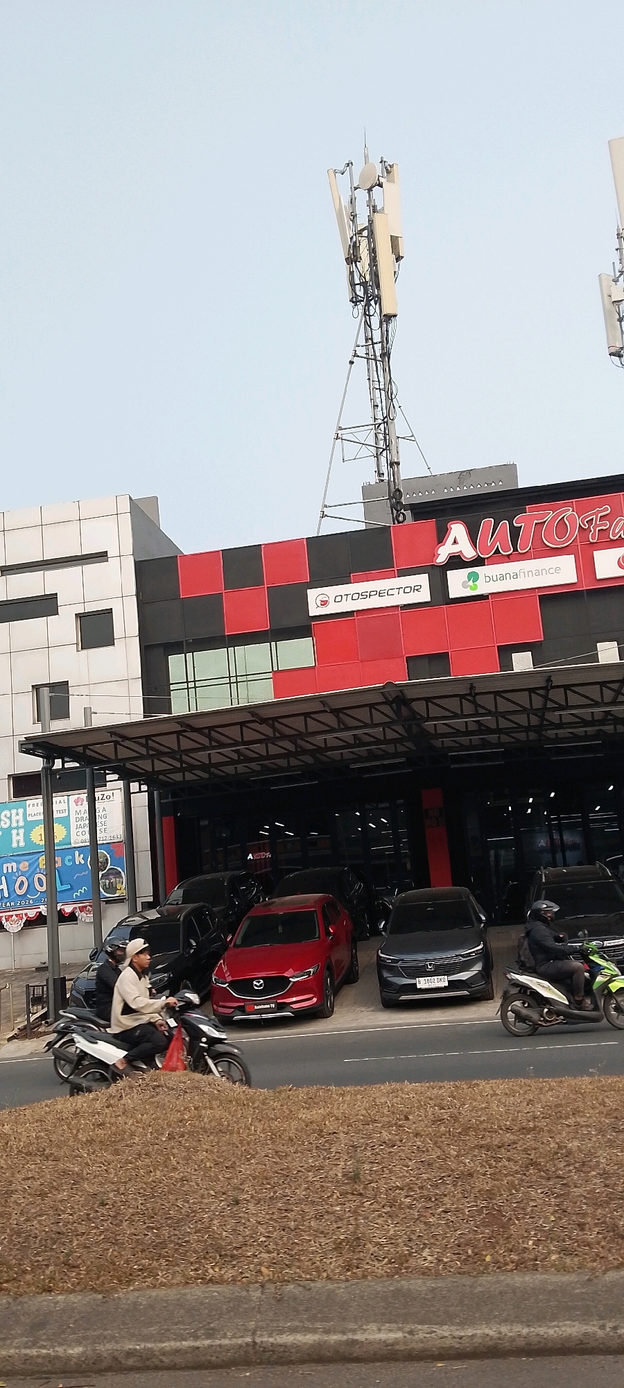 The image depicts a street scene in Gunung Putri, Indonesia, featuring a car dealership and surrounding infrastructure. In the foreground, dry, brown foliage covers a sloping embankment and a concrete curb. A paved road runs horizontally across the lower portion of the image, with two motorcycles and several cars visible.
On the left, a person wearing a light-colored jacket and dark pants rides a white scooter. A red bag is visible on the scooter. Behind the scooter, several dark-colored cars are parked under a covered area. To the right of these cars, a red Mazda SUV is prominently displayed. Further to the right, a dark grey SUV with a license plate B 1002 CKD is parked. Another motorcycle, this one green and white, is being ridden by a person wearing dark clothing and a helmet, to the right of the grey SUV.
The background is dominated by a modern building with a distinctive facade. The building features sections of black and red checkerboard paneling, as well as silver and white panels. A large sign with the words "AUTO F" and a stylized "F" in white lettering against a red background is mounted on the building. Below this sign, smaller logos for "OTOSPECTOR" and "buana finance" are visible. The building is topped by telecommunications towers. To the left of the main building, a section of another building with dark windows is visible. In the far left of the image, a sign with blue lettering reads "FREFRI... TesH H... me HOO".