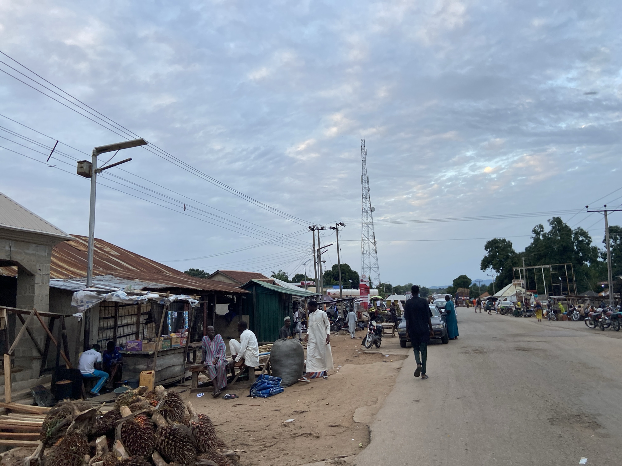 The image depicts a street scene in Mararaba, Nigeria. The road is partially paved and shows signs of wear. A communication tower is visible in the background. Several individuals are present, including men in traditional attire. Some people are standing and conversing near makeshift shops and stalls. Motorbikes are parked along the side of the road. Piles of palm fruits are in the foreground. Overhanging power lines and a street light are also present. The sky is overcast.