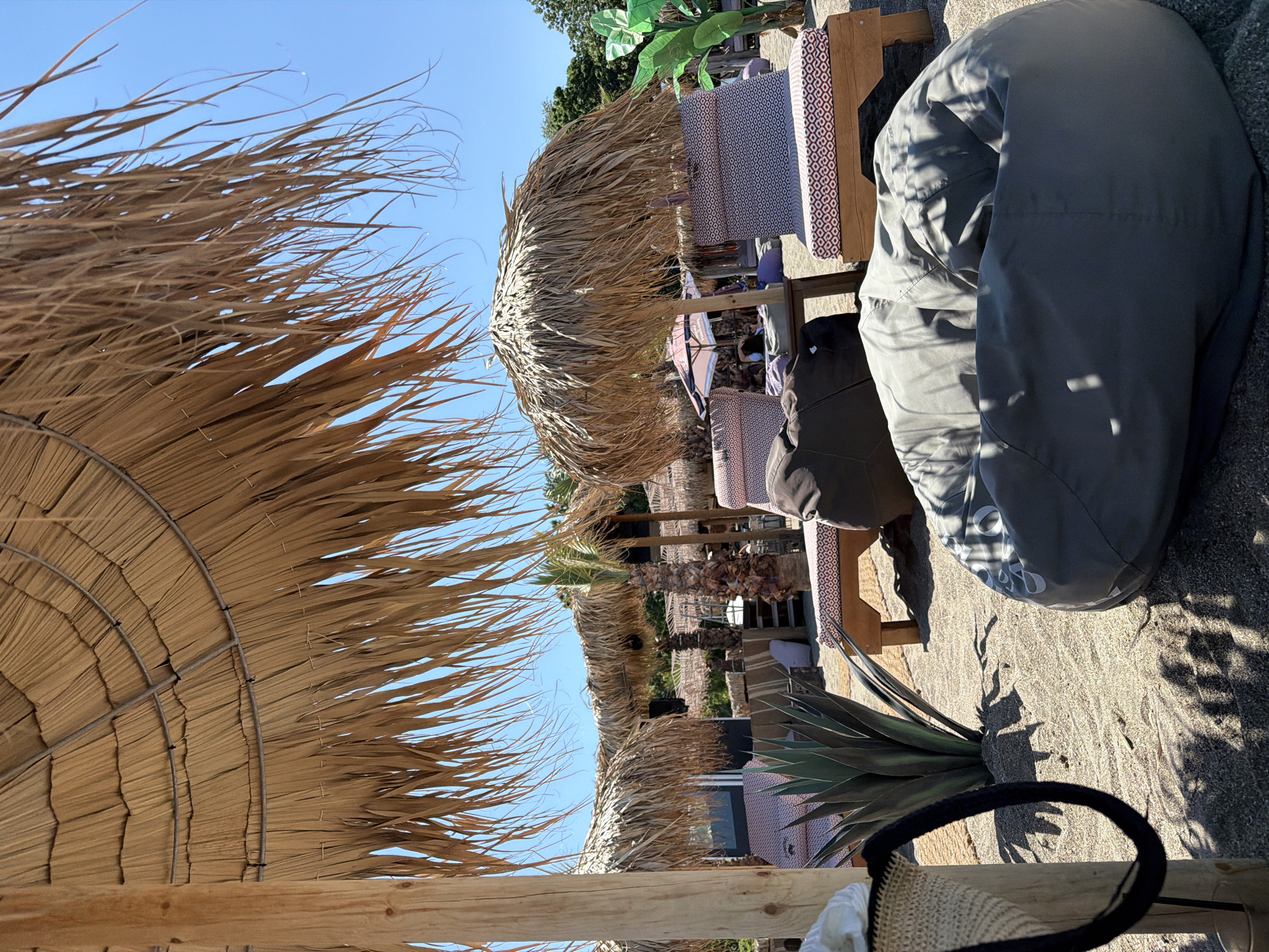 The image depicts a beach club scene in Burgas, Bulgaria, likely on the city's beach or a nearby resort. In the foreground, a large, grey beanbag chair sits on the sand, partially shaded by a thatched umbrella. To the left, a straw bag hangs from a wooden post supporting another thatched umbrella. Behind the beanbag and to the left, an agave plant stands in the sand. Further back, several wooden lounge chairs with red and white patterned cushions are visible, also under thatched umbrellas. A small palm tree is visible behind one of the umbrellas, and in the background, a low-rise building with dark windows is present. The sky is clear and blue, indicating a sunny day. The overall impression is one of a relaxed, outdoor seating area designed for beachgoers.