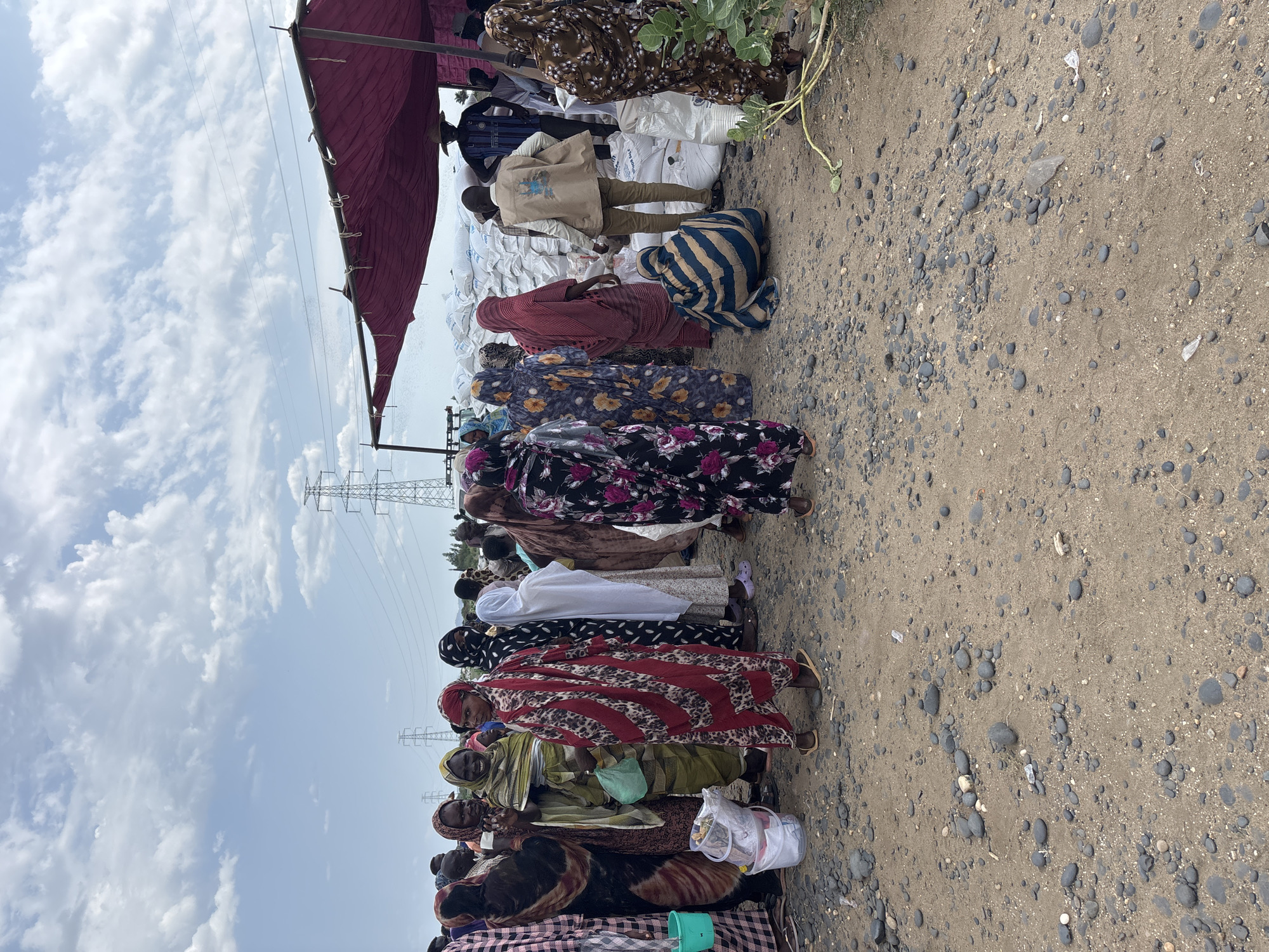 The image depicts a group of people, predominantly women, gathered outdoors in what appears to be a distribution or aid site. The ground is covered with gravel and sand. In the background, several large white sacks are stacked under a burgundy-colored tarp, suggesting they contain supplies. Tall electricity pylons are visible in the distance, and the sky is partly cloudy. Some individuals are wearing colorful traditional clothing, while others are in more neutral attire. One person in the foreground, wearing a vest with a logo, appears to be an aid worker. The scene suggests a humanitarian aid distribution or a market setting where goods are being exchanged. The specific location in Ad-Damazin, Sudan, is not explicitly identifiable from the visual cues alone, but the presence of a large group of people in an outdoor setting with stacked sacks of goods is consistent with aid operations in regions facing hardship.