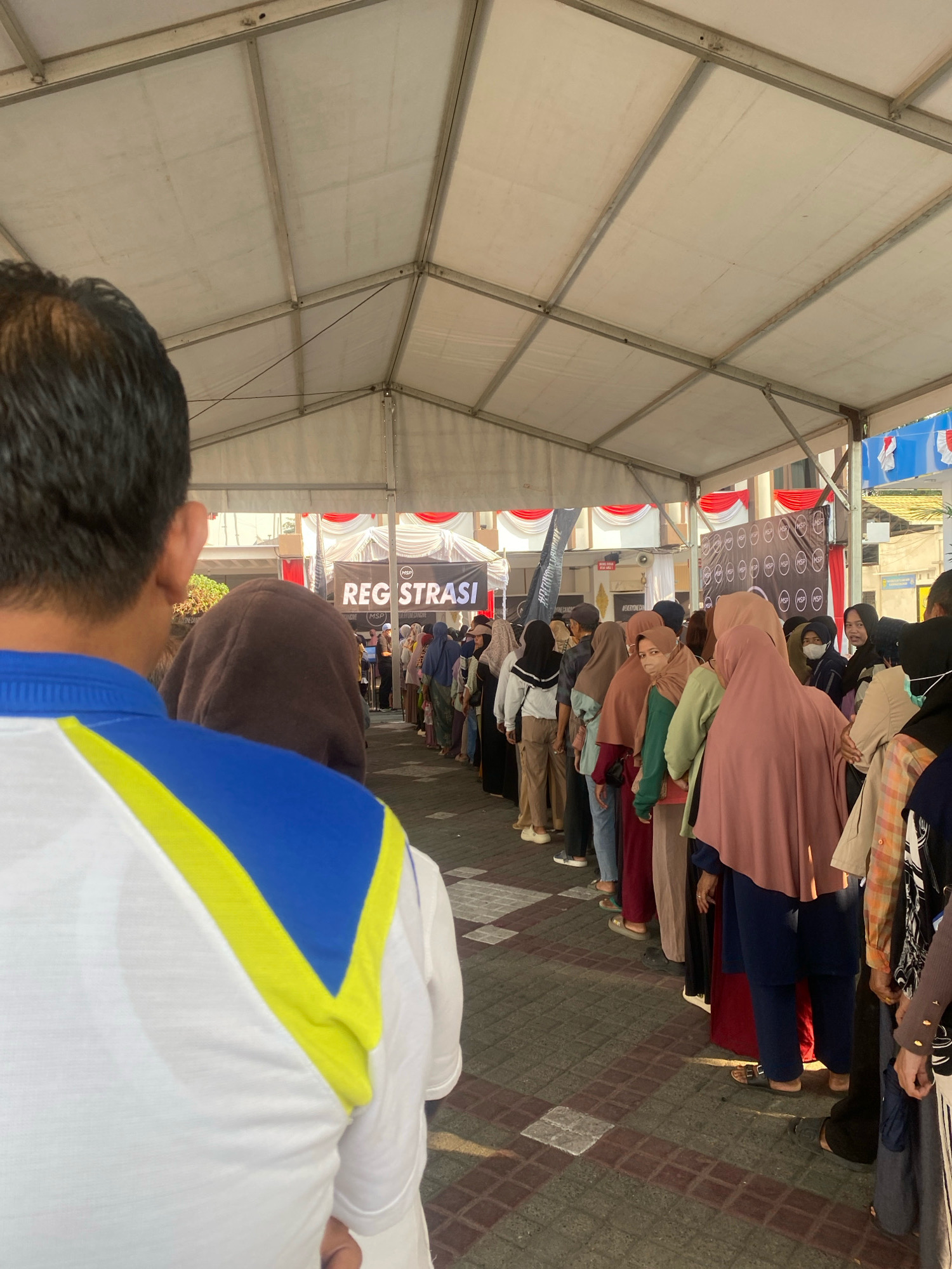 In Banjarmasin, Indonesia, a line of people stands under a white tent. The tent has a metal frame and is open on the sides, revealing buildings and a street outside. A banner displaying "REGISTRASI" (Registration) is visible in the background. Most people in the line are wearing headscarves, indicating it might be a religious or cultural event. The ground is paved with square tiles. The lighting suggests it is daytime.