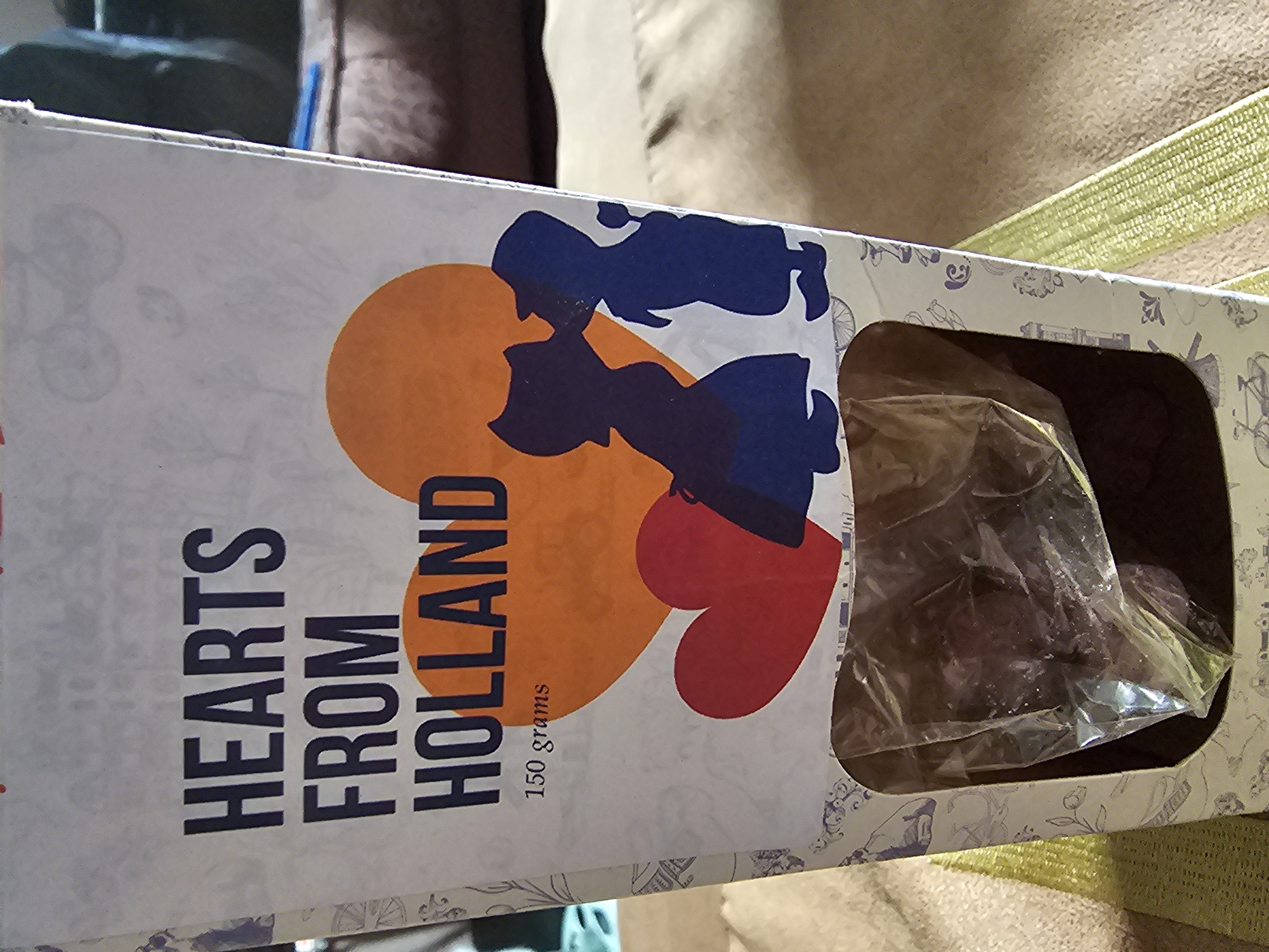 A sideways close-up shows a box of "Hearts from Holland" chocolates, weighing 150 grams. The box is white, and features an orange, red, and blue graphic of a couple. The words "Hearts from Holland" are printed in large, bold, dark-blue letters. An opening in the box displays a clear plastic bag filled with the chocolates. The box is placed on a light-brown or beige surface, possibly a couch or similar furnishing. The background is out of focus, showing darker fabric colors. No people or specific location details (such as Holywell, United Kingdom) are visible in this image.