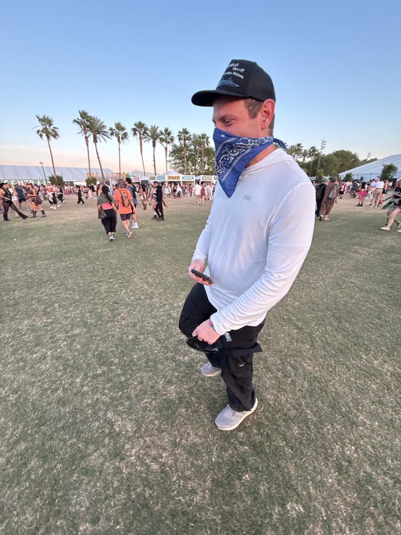 The image depicts a person in the foreground standing on grass in an open area. The person is wearing a black baseball cap, a blue bandana over their face, a light-colored long-sleeved shirt with an "HH" logo, dark pants, and gray shoes. They are holding a black phone in their right hand. In the background, there is a crowd of people and several palm trees. The setting appears to be outdoors, likely at an event or festival. Given the presence of palm trees and the festival-like atmosphere, it is plausible that the location is in Indio, United States, known for hosting events like Coachella.