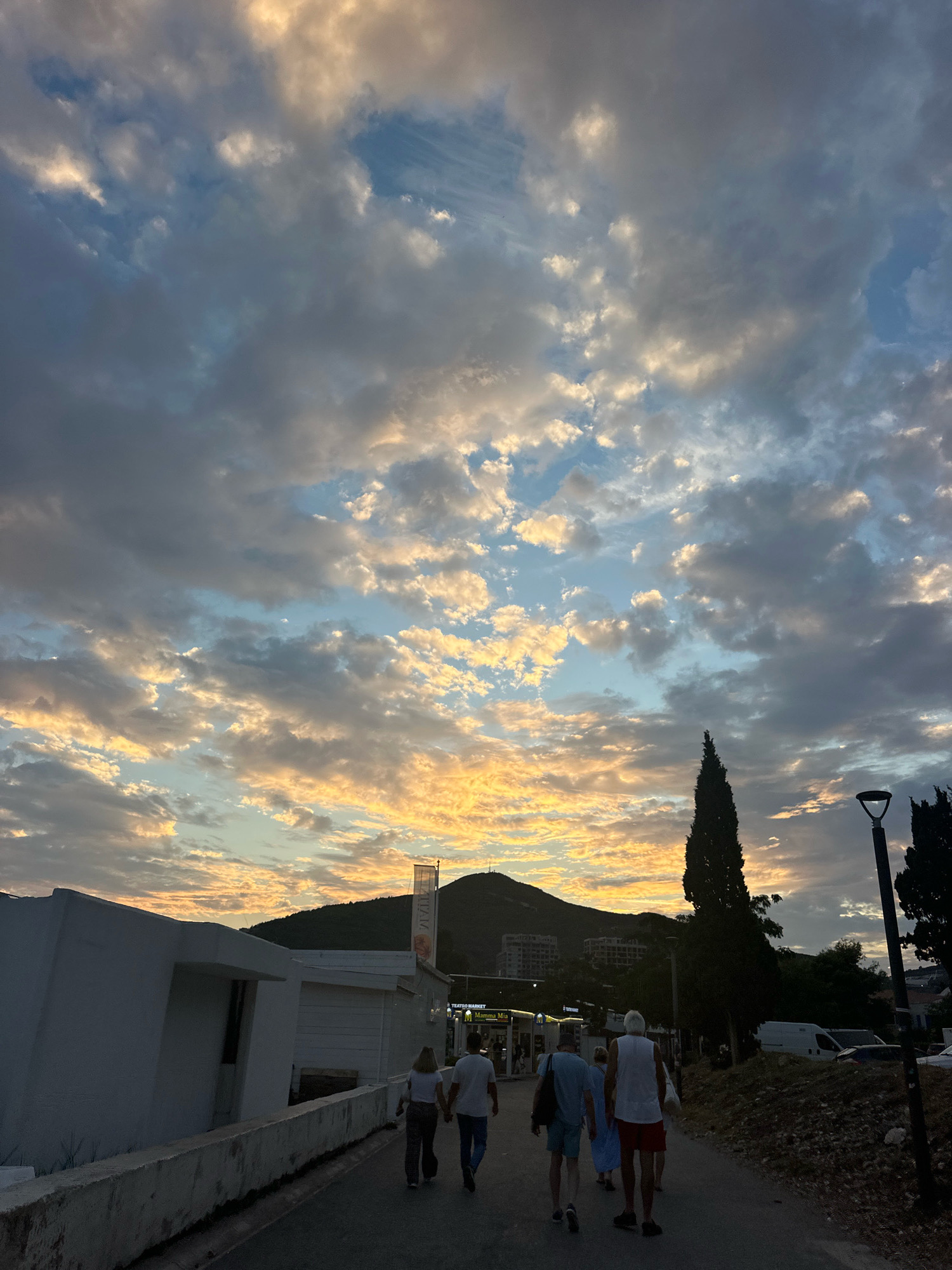 The image depicts a scenic view of Budva, Montenegro, at dusk. The sky is filled with dramatic clouds, illuminated by the setting sun, casting a warm glow of yellow and orange hues. In the foreground, a group of people are walking away from the camera along a paved path. To the left, a white building with a modern architectural design is visible. The path is lined with a low concrete barrier. Further in the background, lush green hills rise, dotted with buildings. A tall, slender evergreen tree stands prominently on the right side of the path, and a modern lamppost is also visible. The overall atmosphere is peaceful and serene, capturing the beauty of a coastal town at twilight.