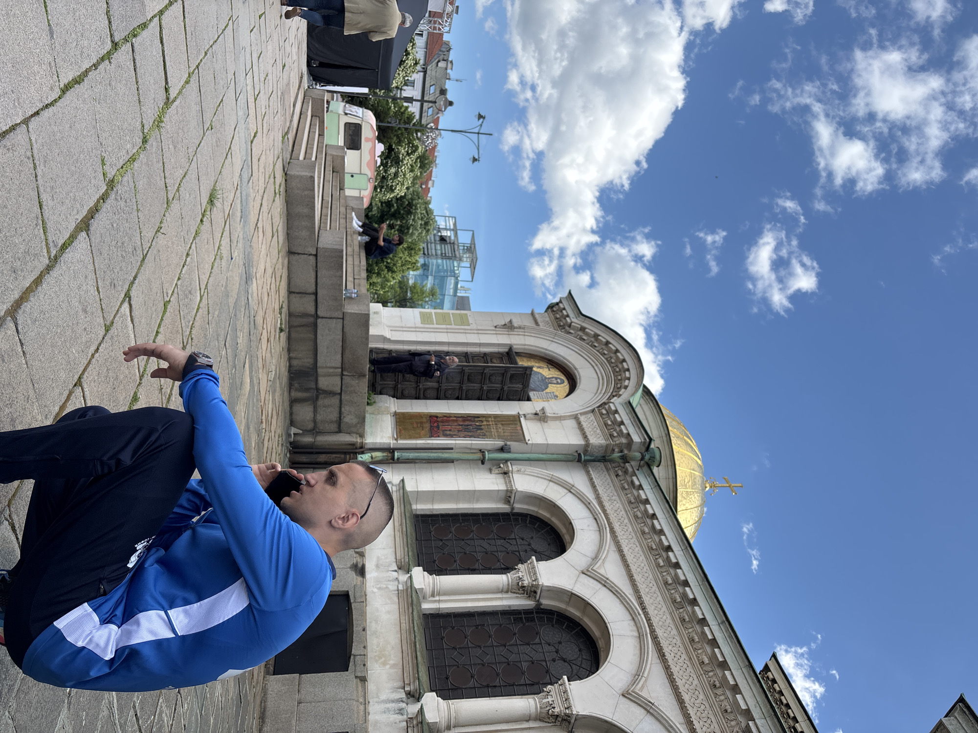 The image shows a man sitting on a stone surface next to the Alexander Nevsky Cathedral in Sofia, Bulgaria. He's wearing a blue long-sleeved shirt with a white stripe and dark pants. He appears to be on a phone call. The Cathedral, a large, ornate building with a gold dome and a cross on top, is partially visible in the background. Above it, there's a clear blue sky with scattered white clouds. Other people, a vehicle, and buildings can be seen in the further background. The foreground features a stone-paved area.