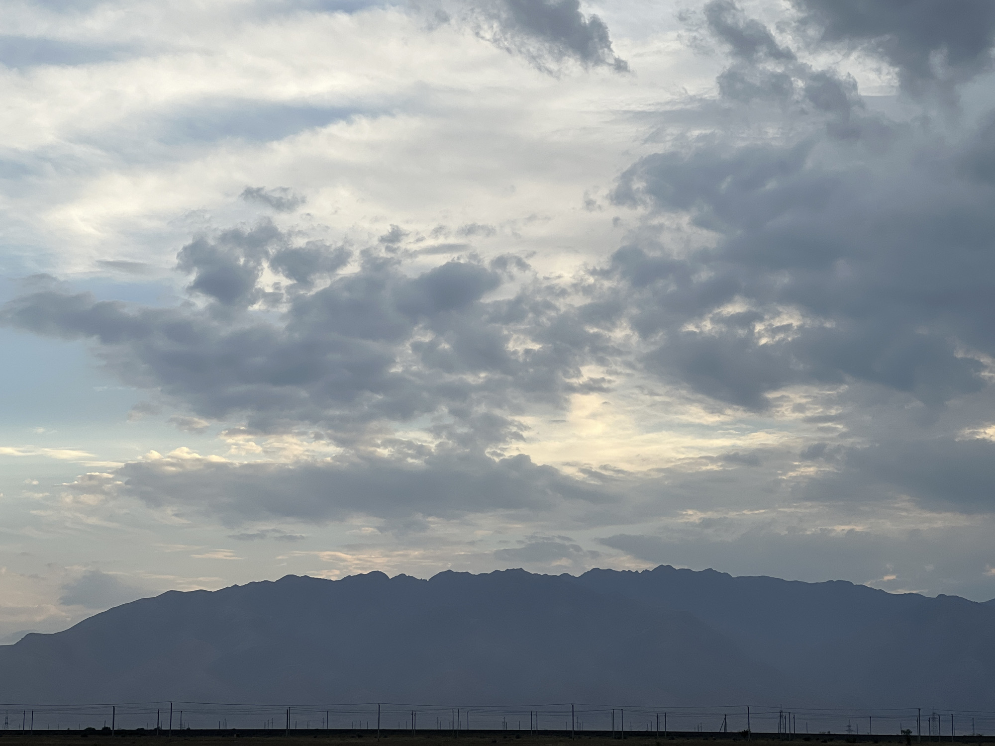 A serene, wide-angle shot captures a mountain range silhouetted against a dramatic sky filled with a mix of dark and light clouds. The mountains, a deep blue-gray, stretch across the horizon, their jagged peaks creating a textured outline. Below the mountains, a flat, arid plain is visible, dotted with numerous vertical poles that suggest a power line or fence. The sky is a gradient of soft blues and whites, with patches of brighter sunlight breaking through the overcast. The overall impression is one of vastness and natural beauty. The scene is located in the vicinity of Ақши, Kazakhstan. There are no people or animals visible in the image.