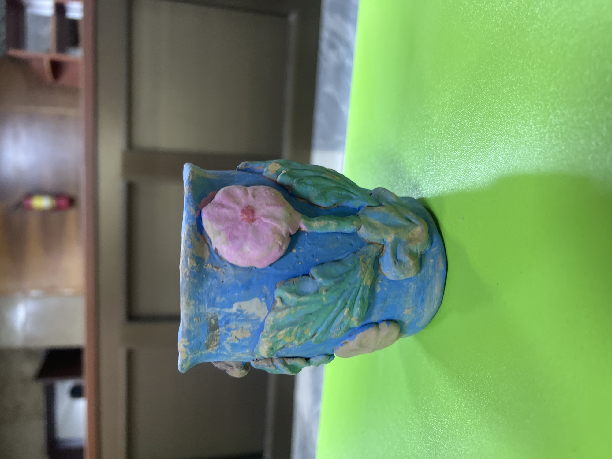 A close-up, eye-level shot shows a handmade blue clay pot with raised pink and green floral and leaf designs. The pot is positioned on a bright green surface. In the background, a wooden structure with shelves is visible, and a red fire extinguisher hangs on the wall. The scene is indoors.