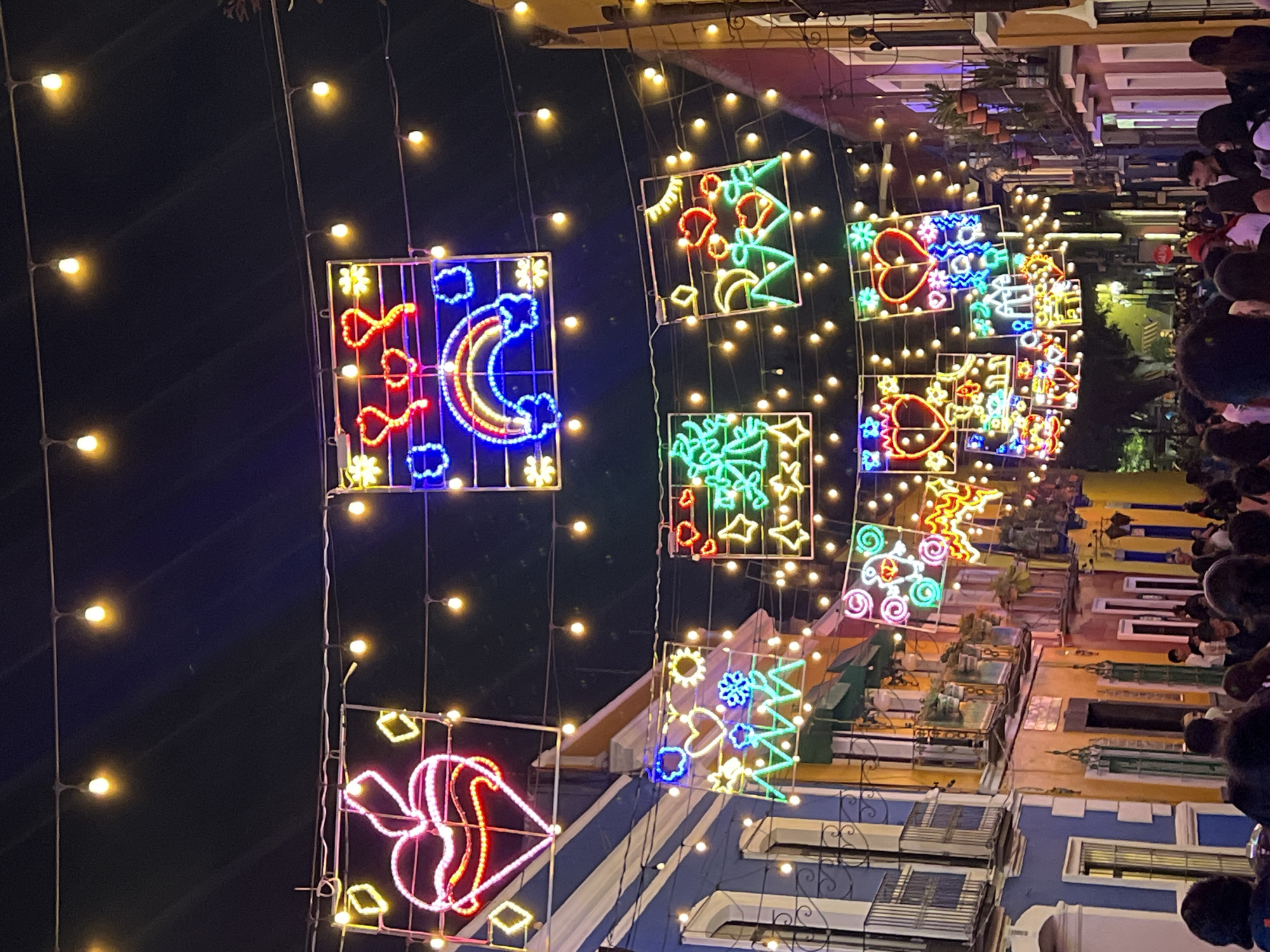 The image shows a street in an unidentified city at night, decorated with strings of lights and illuminated, framed figures suspended above the street. The figures contain images of hearts, moons, clouds, snowflakes, and other festive shapes. The buildings lining the street are mostly painted blue and yellow. People are gathered on the sidewalk to view the decorations.