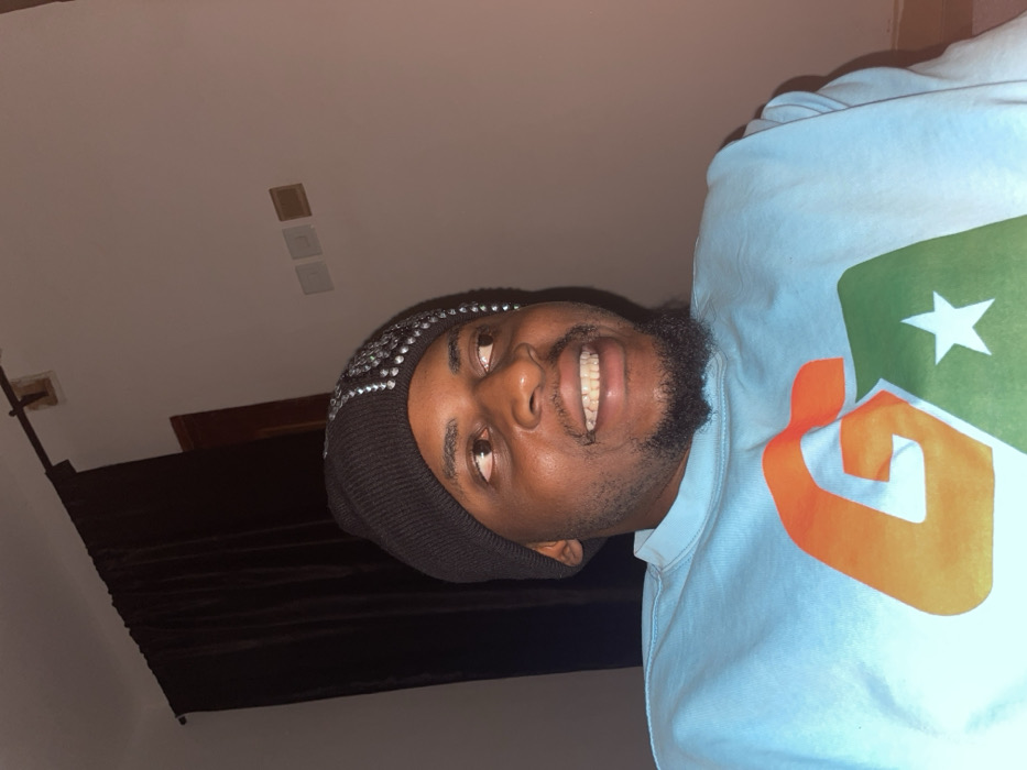 The scene depicts a male individual positioned indoors, located in Ube, Nigeria. He has a dark complexion, a short beard, and is wearing a black knit beanie embellished with silver reflective studs on the right side. His attire includes a light blue long-sleeved sweatshirt featuring a prominent graphic of an orange 'G' partially overlaid by a green shape containing a white star. The individual's head is turned slightly to the right, with his eyes directed towards the right side of the frame and upward. He exhibits a wide smile, revealing white teeth. The background features a plain light-colored wall. Dark brown curtains hang from a rod on the left side of the frame. On the wall to the right, three rectangular elements, consistent with electrical outlets or switches, are visible in white, light brown, and darker brown finishes.