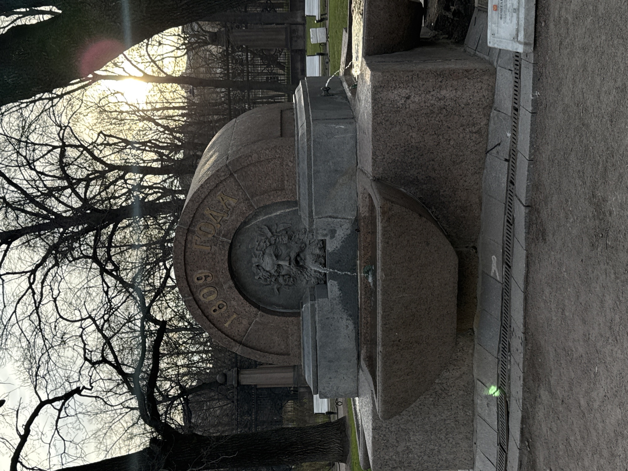 The image shows a drinking fountain in Saint Petersburg, Russia. The fountain is made of granite and features a semi-circular basin with a mask from which water flows. Inscribed on the upper part of the basin is the year "1809". The fountain is surrounded by bare trees. In the background there is a wrought iron fence and a grassy area with benches. The sun is shining through the trees, creating a lens flare effect. The image is taken at ground level and is rotated counter-clockwise.