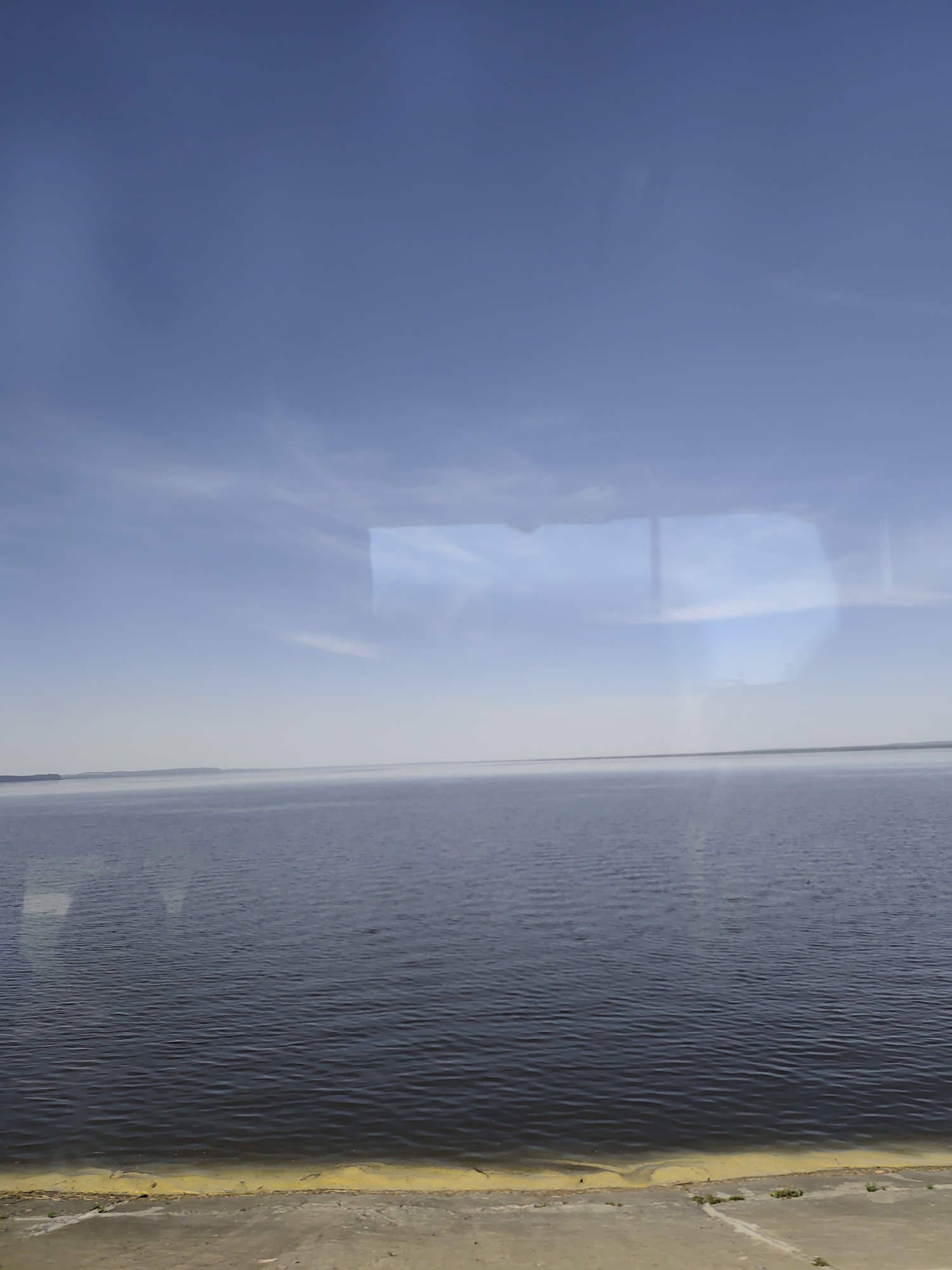 The image depicts a body of water, likely a river or lake, under a blue sky with some clouds. The water appears calm with slight ripples. In the foreground, there is a concrete embankment with a yellow line marking the edge. Small patches of vegetation are visible along the embankment. The location is implied to be Chaikovsky, Russia. There are no people or specific objects visible in the image beyond the natural elements and the man-made embankment. No actions or interactions are depicted.