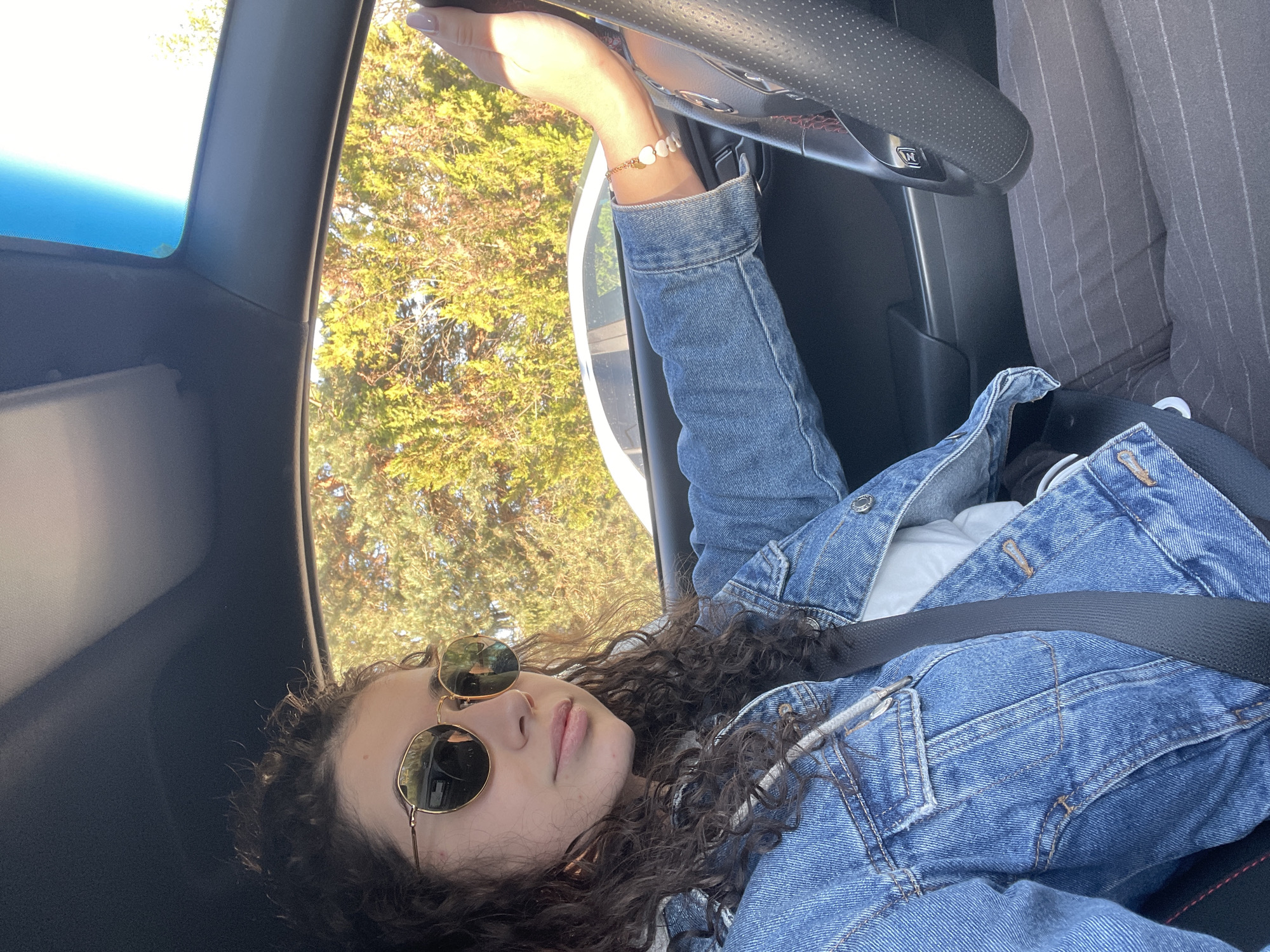 A young individual with curly dark hair and reflective round sunglasses is reclining in the passenger seat of a car, facing towards the viewer. They are wearing a blue denim jacket over a white top, secured by a black seatbelt across their chest. A bracelet with light-colored, possibly heart-shaped, beads and a small golden charm is visible on their left wrist. Their left arm is extended upwards, resting on the black, textured steering wheel, which features a metallic logo that appears to be a stylized 'W' or 'M'.
The car's interior shows a pinstriped seat beside the individual, parts of the dashboard, and an A-pillar. Through the car windows, lush green and yellow-green trees are brightly lit by sunlight, indicating a clear, sunny daytime. A light blue tint is visible at the top of the windshield or sunroof. The individual's expression is neutral, with their mouth slightly open, though their eyes are obscured by the sunglasses. The car appears to be left-hand drive, fitting the context of Sofia, Bulgaria.