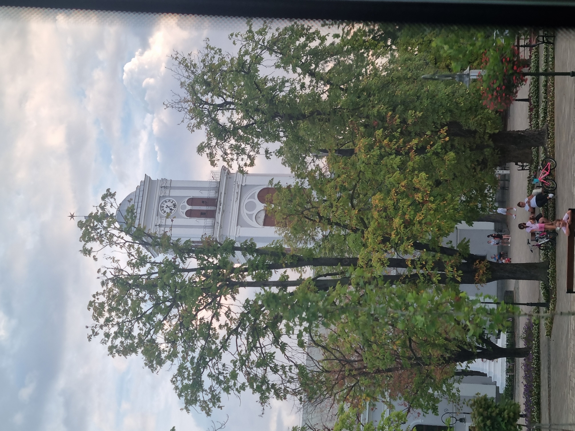 The image shows a view of the clock tower of the Salonta Orthodox Cathedral in Salonta, Romania. The clock tower is tall and white with arched windows and a star on top. Large trees with green leaves are in the foreground, partially obscuring the view of the cathedral. In the background, there are people walking and sitting in a park area. Some children are playing with a pink bicycle. The sky is cloudy with patches of blue.