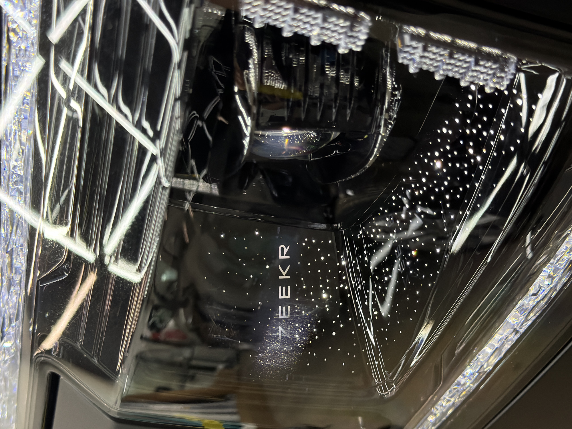 The image shows a close-up of a car headlight. The headlight's interior is visible, revealing LED light patterns, a projector lens, and the car brand "ZEEKR" printed vertically. The headlight assembly is encased in clear plastic, with internal reflective elements and light diffusion patterns visible. The background is out of focus, hinting at a car interior and external environment. No specific location in Krasnoyarsk, Russia, can be determined from the image. There are no people visible, and no specific actions or interactions are occurring within the frame.
