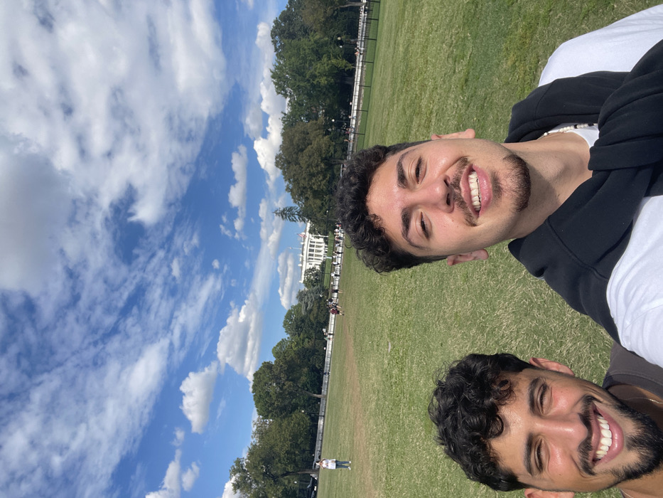 Two young adult males are featured in a selfie-style photograph. The male on the right, centrally positioned and smiling broadly with visible teeth, has dark curly hair, a mustache, and a chin-strap beard. He wears a white t-shirt with a black sweatshirt draped over his shoulders and a white beaded necklace. The male on the left is partially framed, smiling with visible teeth, possessing dark curly hair and a beard, and wearing a dark t-shirt.
They are situated on a large, vibrant green grass lawn. In the midground, a dark security fence delineates the boundary of the lawn, behind which numerous indistinct individuals are visible. Further in the background, a dense line of green trees partially frames the iconic white neoclassical building of the White House, distinguished by its columns and a flag flying on the roof. The sky above is bright blue with scattered white cumulus clouds.
The image was captured in Washington, D.C., United States, with the subjects positioned on a public park area, likely the Ellipse, facing the South Lawn and South Portico of the White House. The scene depicts a clear, sunny day.