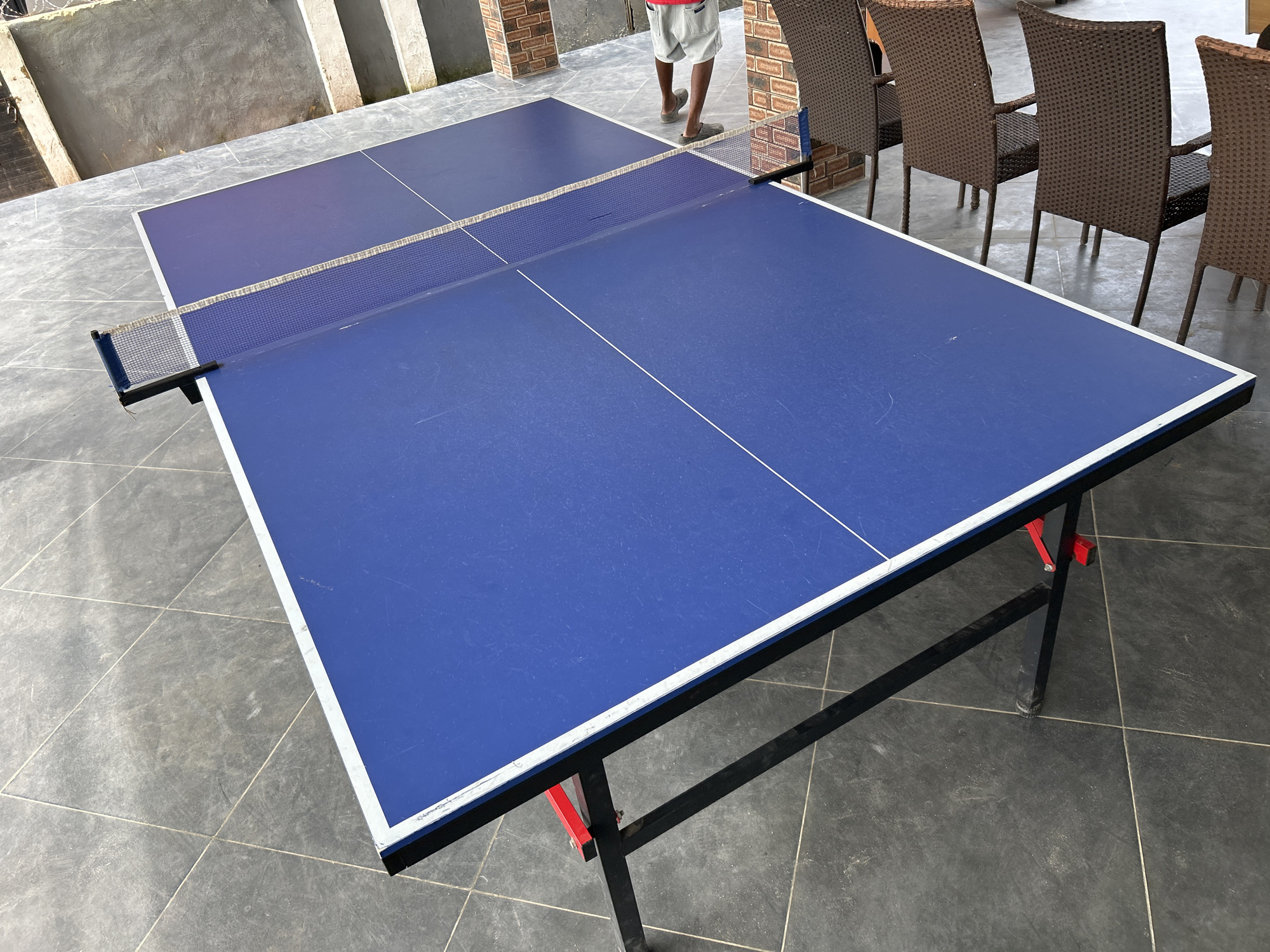 An outdoor ping pong table is set up on a tiled patio area in front of a building. The ping pong table is blue with a white border and a net stretched across the middle. A person wearing a red shirt and grey shorts stands behind the table, facing away from the camera. To the right of the table are several brown wicker chairs. The background shows a building with brick pillars and a grey concrete wall. The ground is covered in dark grey square tiles.