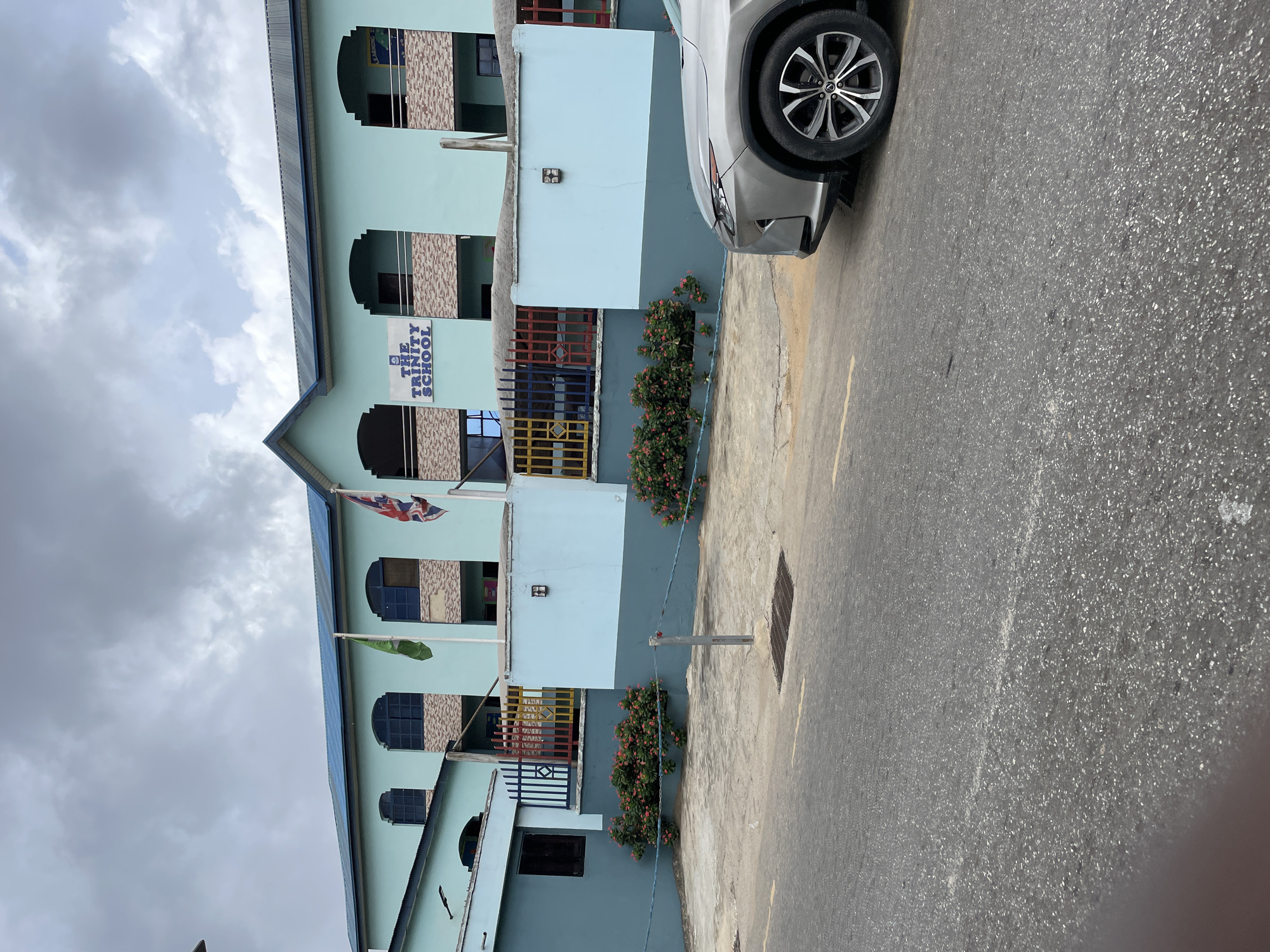 The image shows a multi-story building identified as "The Trinity School," likely located in Ikeja, Nigeria, based on architectural style and the presence of a car bearing a Nigerian license plate. The building has a light blue exterior with dark-framed windows and balconies decorated with colorful railings. A British flag and another green flag are visible. A silver car is parked in front of the building, partially obstructing the view. The street in front of the building is paved, with a manhole cover visible. The sky is overcast.