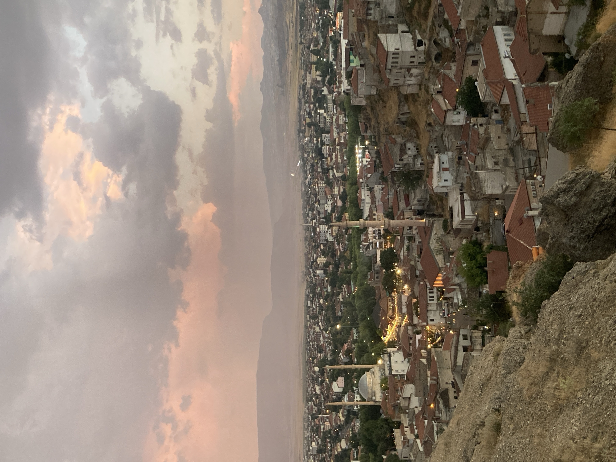 The image is a high-angle, wide shot of Avanos İlçe Merkezi, Turkey, at dusk. The sky is filled with dramatic clouds tinged with pink and orange from the setting sun. In the foreground, the rocky terrain slopes down towards a city composed of densely packed buildings with red tile roofs. Several mosques with minarets are visible, including a prominent one with a large dome and two slender minarets in the mid-ground. The streets are illuminated by streetlights, creating a warm glow across the urban landscape. Distant mountains form the backdrop, shrouded in a haze. No people are visible in the image. The overall impression is a serene and picturesque view of the city at twilight.