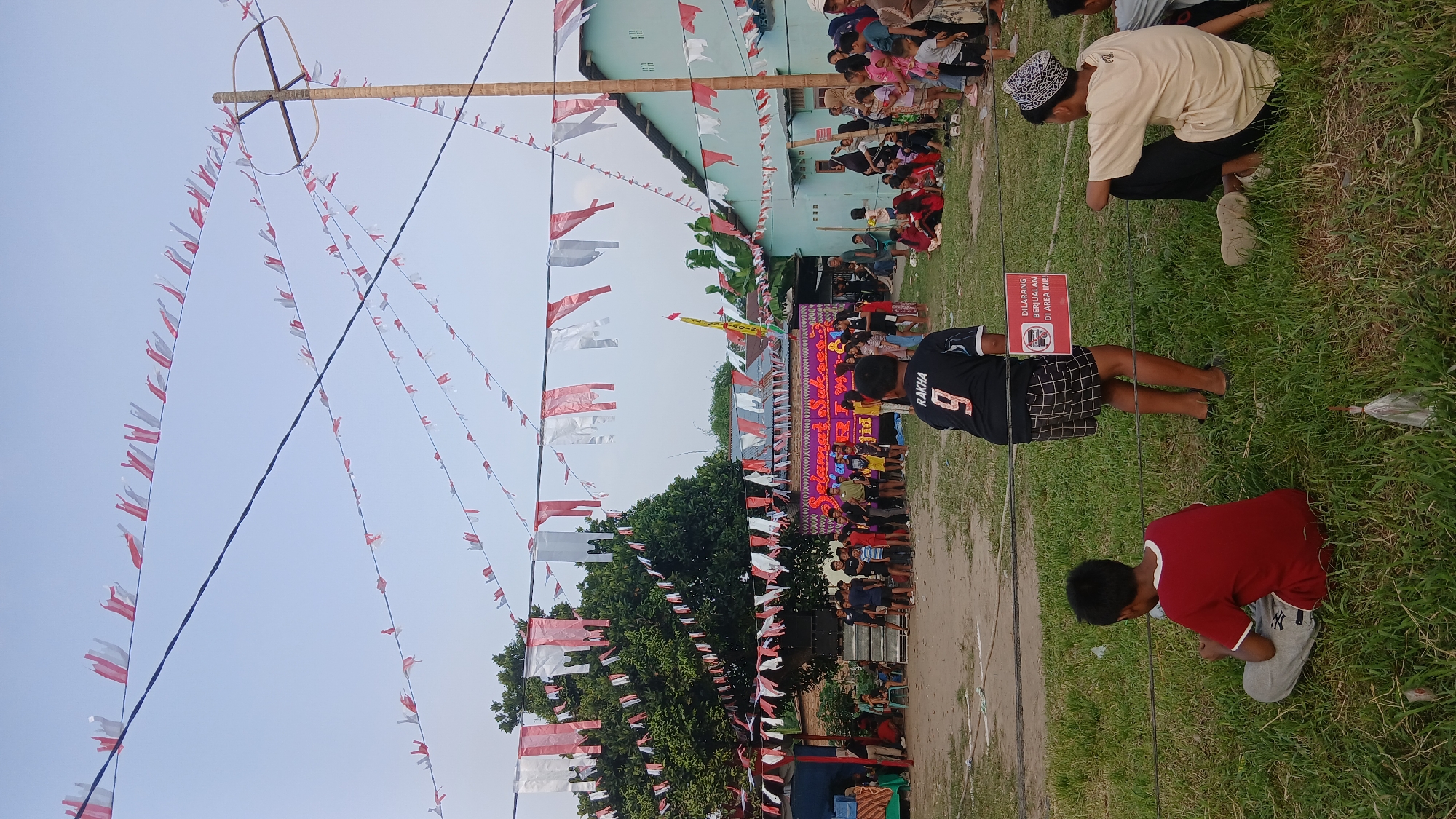 The image depicts an outdoor event in Tanjung Gusta, Indonesia, likely a community gathering or celebration. The scene is decorated with numerous red and white Indonesian flags strung across wires overhead, creating a festive atmosphere. In the background, a building with light blue walls and a corrugated metal roof is visible, along with several trees. A stage area is set up, adorned with a banner that reads "SELAMAT SUKSES" (Congratulations) and other text. A group of people, including children and adults, are gathered in front of and around the stage, some standing and some seated. A young boy wearing a red shirt and grey pants is sitting on the grass in the foreground, facing away from the camera. Behind him, a person in a black jersey with the number 9 is standing and looking towards the stage. A red and white sign is attached to a wire in front of the person in the black jersey, displaying a symbol of a bus and text that translates to "PROHIBITED TO SELL IN THIS AREA !!!". The ground is covered with grass and some bare earth. The overall lighting suggests it is late afternoon or early evening.