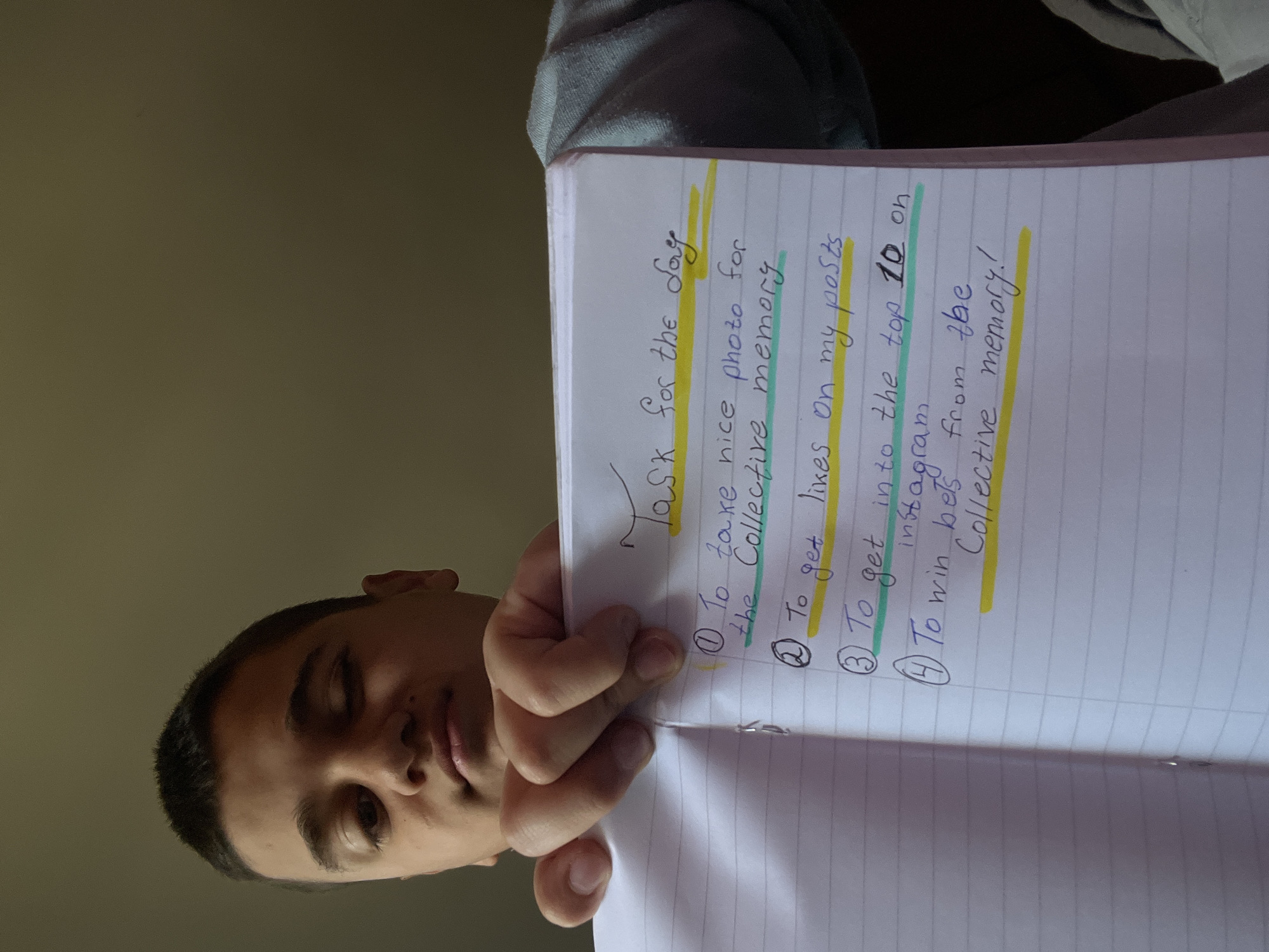 A close-up, low-light shot depicts a person holding an open, lined notebook, presenting a handwritten list. The person, whose head and right hand are visible in the lower left of the frame, has short, dark hair and appears to be looking down at the notebook with a neutral expression. The background is a plain, dark, olive-green color, suggesting an indoor setting with subdued lighting, possibly in Lozarevo, Bulgaria.
The notebook, held vertically, contains a list titled "Task for the Day," written in blue ink with yellow highlighting. The list comprises four numbered items, also in blue ink, with some words highlighted in yellow or green.
The visible text reads:
"Task for the Day
① To take nice photo for the Collective memory
② To get likes on my posts
③ To get into the top 10 on instagram
④ To win bets from the Collective memory!"