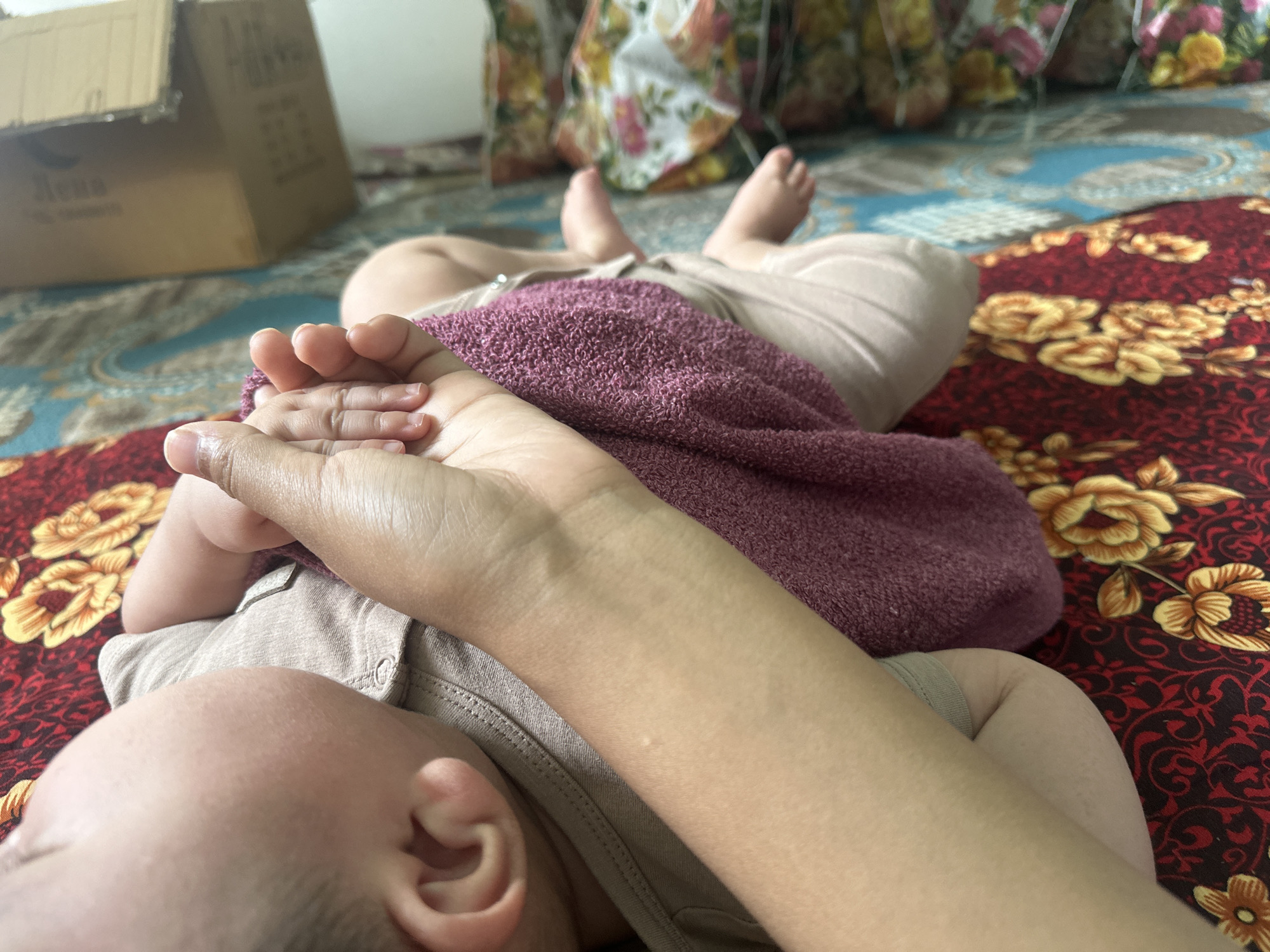 An overhead, close-up shot shows a person's arms and a baby's body lying on a red and yellow floral patterned blanket. The person's right arm is extended across the baby's torso, which is covered by a purple towel. The baby's legs are visible at the top of the frame, with their feet pointing upwards. A cardboard box is visible in the upper left quadrant of the image. The scene is indoors, on a bed with patterned sheets, possibly in a residential setting in Тайгараев, Kyrgyzstan.