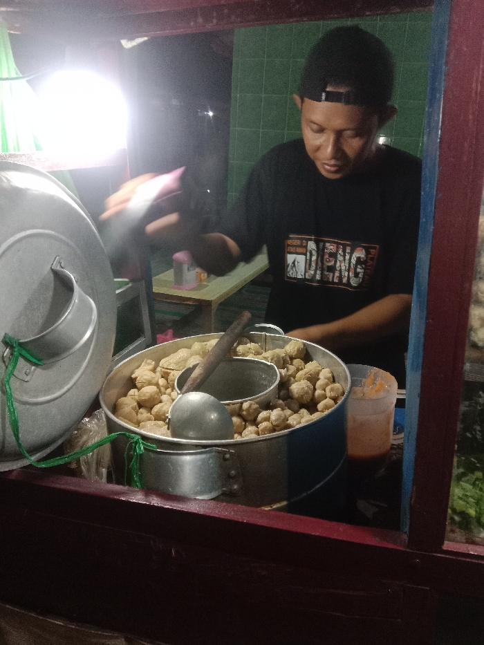 At night in Turunsih, Indonesia, a male vendor operates a street food stall. The vendor, wearing a black t-shirt and a backward black baseball cap, stands behind a counter. He is actively attending to a large aluminum pot containing numerous light-brown, round food items, likely meatballs (bakso). A smaller aluminum container, acting as a steamer insert, also holds similar items within the larger pot. A metal ladle rests among the food. To the right of the pot, a clear plastic cup holds a reddish-orange sauce. A large, domed aluminum lid, secured with a green string, is visible on the left. A bright, circular light source illuminates the scene from the left. In the background, a light-colored wooden table is visible, and behind it, a wall with green tiles. The foreground shows parts of a wooden stall structure painted dark red and blue. The vendor's right hand is blurred, indicating motion.