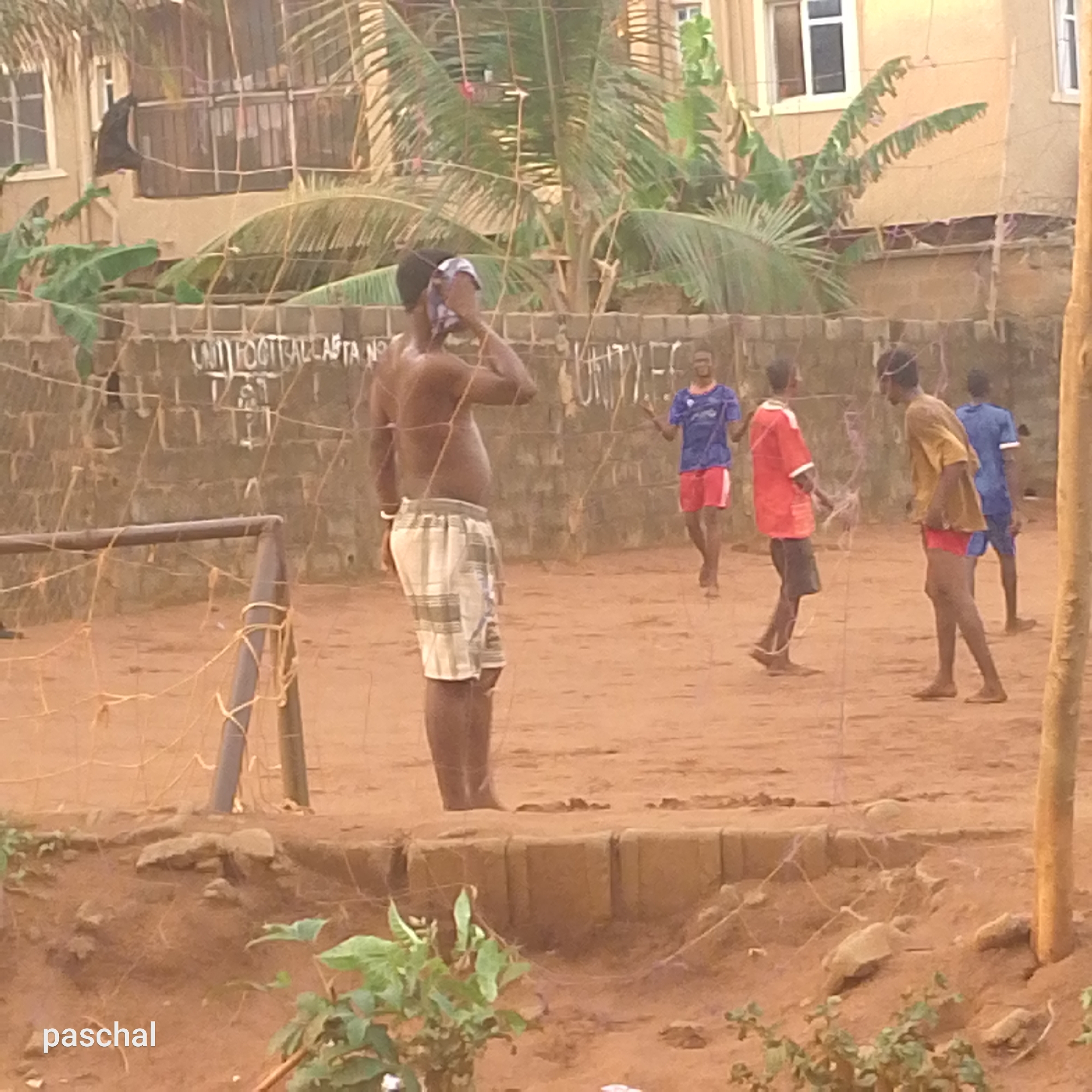 The scene depicts an outdoor soccer game in Ukwu-Abwa, Nigeria. The setting is a dirt field enclosed by a brick wall and dilapidated netting. In the foreground, a bare-chested man in plaid shorts dabs his face. Four young men, dressed in mismatched sports attire, are playing soccer in the middle ground. Buildings and palm trees are visible in the background. A soccer goal is positioned on the left side. The ground is uneven with vegetation in the foreground. The name "paschal" is in the bottom left corner. The wall behind the field is tagged with the phrases "UNIT FOOTBALL ALLSTA NO" and "UNITY FC."