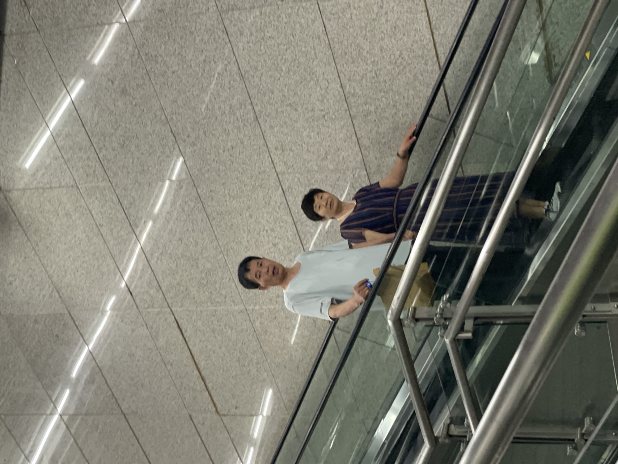 The image shows a man and a woman riding an escalator, likely in an indoor public space in Yuzhong District, China. The man is in the foreground, wearing a light blue short-sleeved shirt and khaki pants. He is holding onto the escalator handrail. The woman is behind him, wearing a striped dress. The escalator has clear glass sides with metal railings. The background is a wall with linear lighting, suggesting the space is a modern building like a subway station.