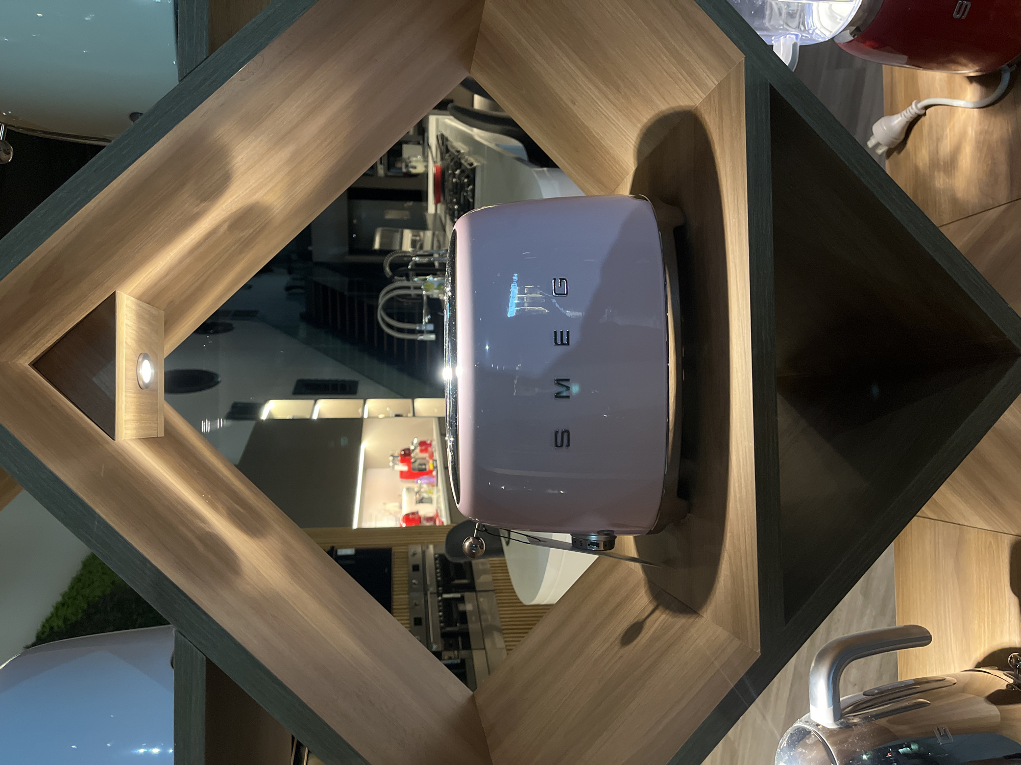 A tilted, indoor shot shows a pink SMEG toaster centered inside a wooden diamond-shaped display case. The toaster has the SMEG logo vertically emblazoned on its front. Through the toaster's reflective surface and the case's open sides, there is a partial view of a kitchen showroom interior, including other appliances and display fixtures. To the lower left, a portion of a white appliance is visible. To the upper right, a red appliance with a white cord is seen. The case is constructed from light brown wood. The scene appears to be a retail or exhibition space. The location cannot be confirmed as Buenos Aires, Argentina, from this single image.