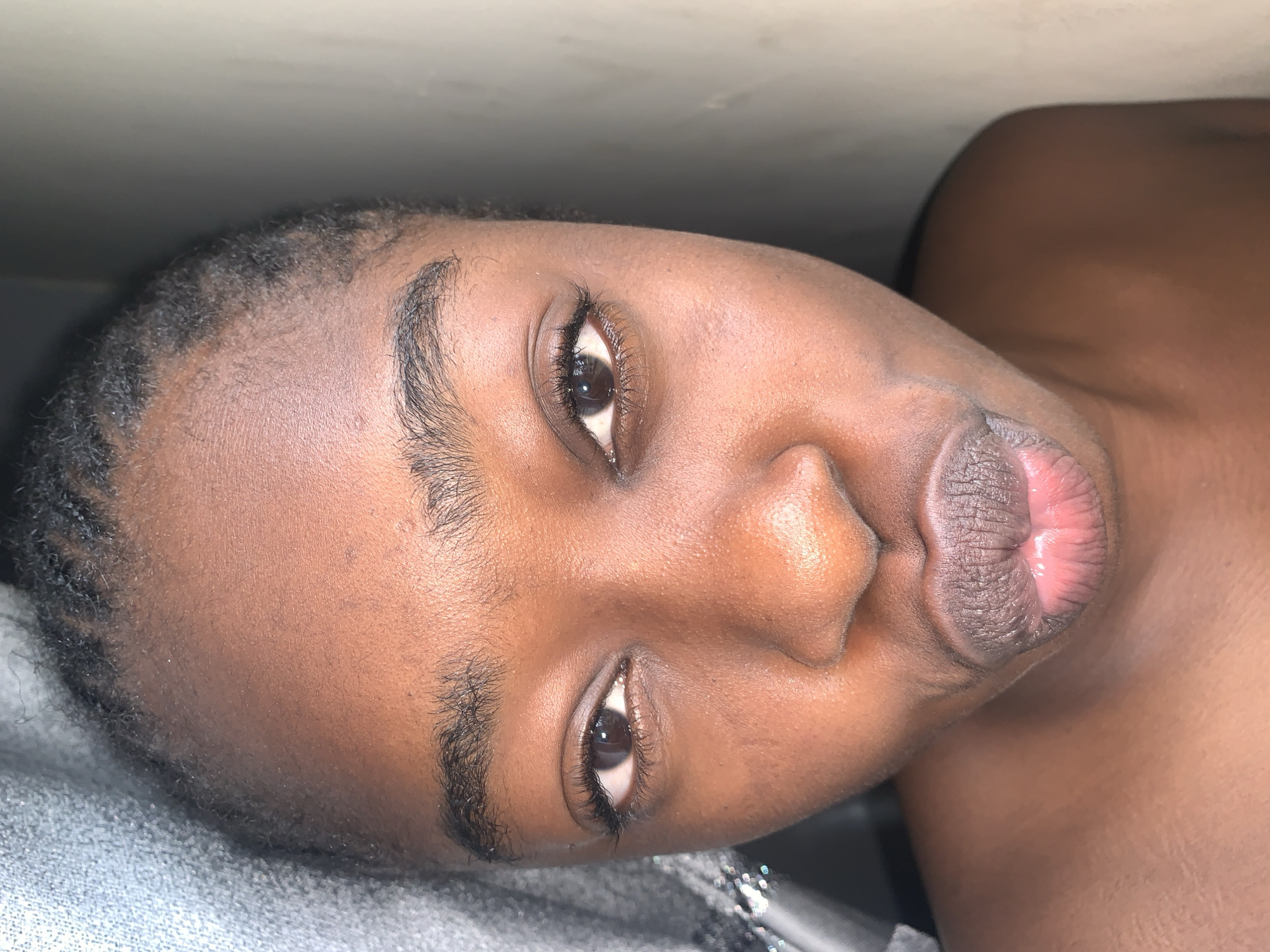 The image is a close-up portrait of a young Black person, likely a female, facing forward and making a kissing gesture. The subject's face fills most of the frame, with the top of their head and shoulders visible at the bottom. They have dark skin, dark eyes, and dark hair styled in braids pulled back from their face. Their lips are slightly parted, with the lower lip extended forward. They are wearing what appears to be light-colored or nude-toned lipstick, which creates a noticeable sheen on their lips.
The lighting suggests an indoor environment, possibly near a window, as indicated by the soft shadows and the slightly uneven illumination on the face. To the left of the subject, a textured, grey fabric is visible, which could be a curtain or clothing. To the right, a plain, light-colored wall is visible.
The image does not contain any elements that would allow for the specific identification of the location within Rafin Par, Nigeria. The scene is devoid of any recognizable landmarks, street signs, or environmental cues that are characteristic of a particular place within the city. The focus is entirely on the subject's face and the immediate, indistinct background.