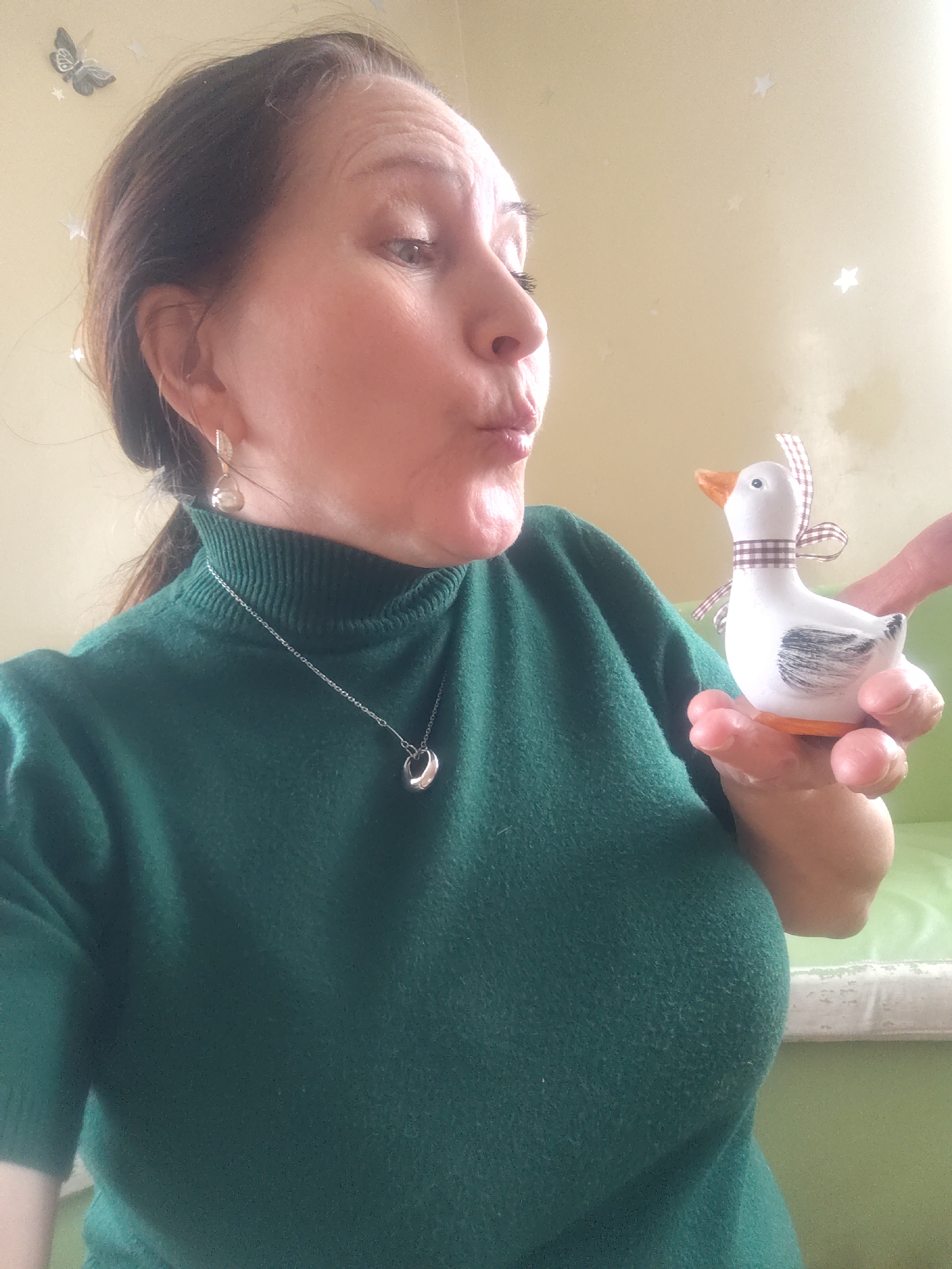 The image shows a woman with fair skin and dark hair pulled back into a ponytail, posing with a small decorative duck. She is wearing a green turtleneck sweater, silver earrings, and a silver necklace with a circular pendant. Her lips are pursed as if she is about to kiss the duck.
The duck is white with a brown beak and brown accents, including a brown-and-white checkered ribbon around its neck. It is held in the woman's right hand. Another hand appears in the background, seemingly holding the duck steady.
The background features a light yellow wall adorned with butterfly and star-shaped decorations. A green surface with a white edge is visible in the lower right corner, suggesting a couch or bed.
There is no information in the image to confirm a location in the city of Sarapul, Russia.