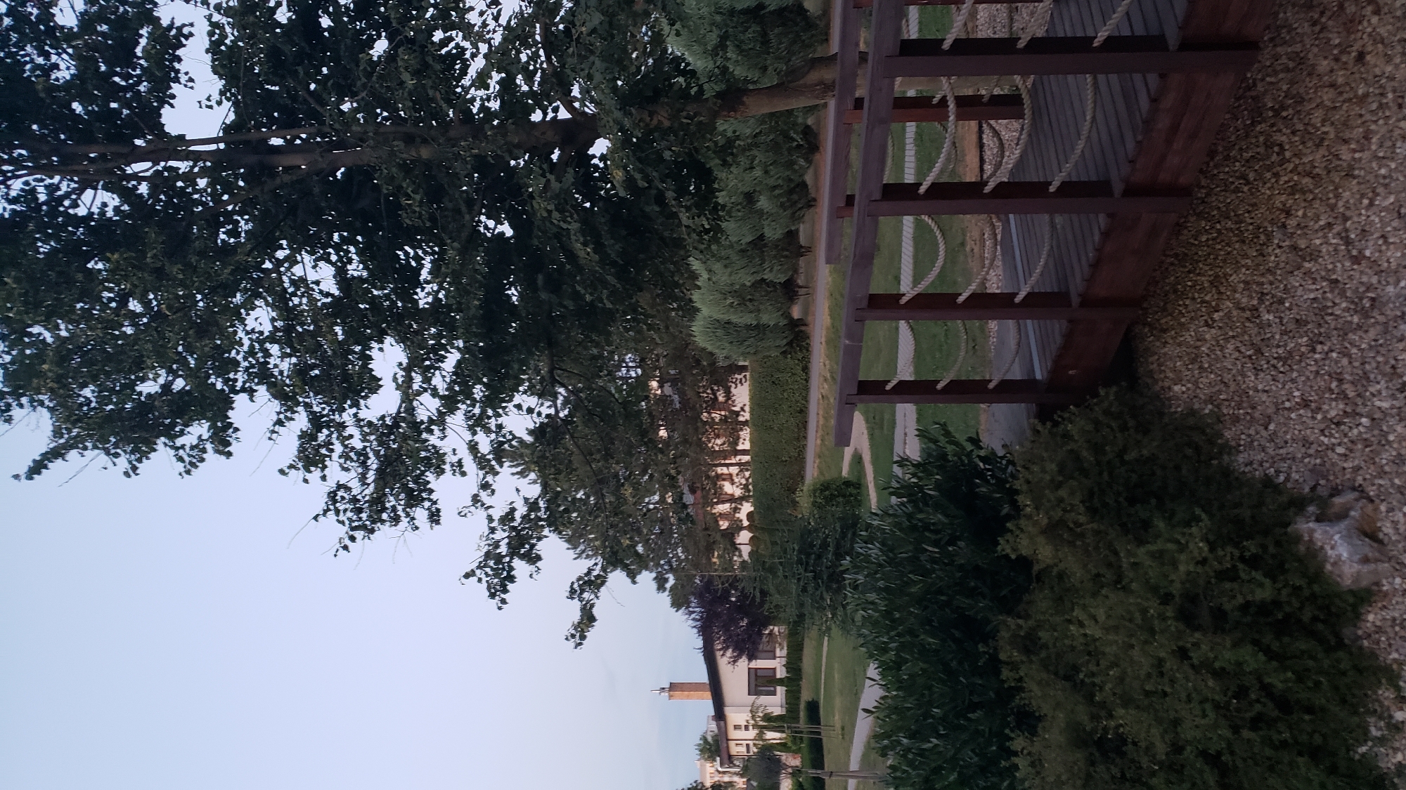 The image depicts a park-like setting with a wooden bridge in the foreground. The bridge has wooden railings with rope detailing and a planked walkway. Beyond the bridge, a grassy area is visible, interspersed with various types of trees and shrubs, including manicured bushes and a large, mature tree. In the background, a white building with a red-tiled roof and a prominent chimney can be seen, along with other structures and dense foliage. The sky is a pale, overcast blue, suggesting it is either early morning or late evening. The scene appears to be a peaceful, outdoor space within a developed area. This location is the Spa hotel Branisko in Prešov, Slovakia, a popular wellness destination known for its tranquil atmosphere and natural surroundings.