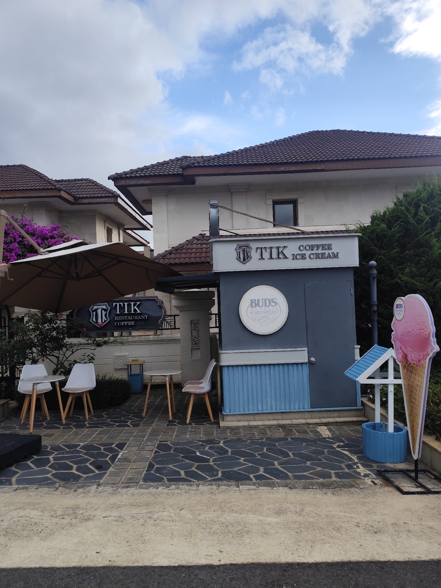 The image shows an outdoor seating area for a cafe and ice cream shop in Lâm Viên - Đà Lạt, Vietnam. A large beige umbrella shades a circular table with two white chairs. To the right, two more white chairs are arranged around a small table. A sign above the outdoor seating reads "TIK RESTAURANT COFFEE". A grey kiosk-style building is visible behind the seating area, with a prominent sign that says "TIK COFFEE ICE CREAM". A circular sign on the kiosk reads "BUDS ICE CREAM OF SAN FRANCISCO". To the right of the kiosk is a large cutout of a pink ice cream cone. The ground is paved with dark hexagonal and rectangular stones. In the background, there are multi-story houses with tiled roofs and lush green trees. The sky is partly cloudy.