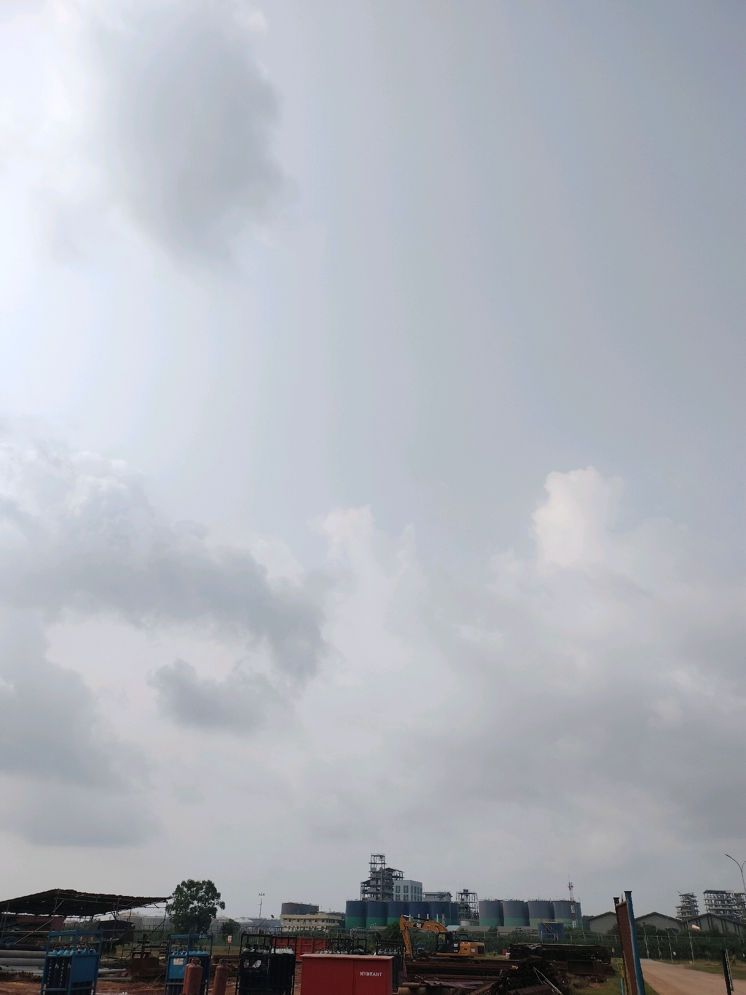 The image shows an industrial area under a cloudy sky, likely located in Batam, Indonesia. In the foreground, there are several industrial tanks, metal scaffolding, construction materials, and a red fire hydrant. A yellow excavator is partially visible in the middle ground, next to piles of scrap metal and other construction debris. Further in the background, large cylindrical storage tanks and industrial buildings are prominent. The overall impression is one of ongoing industrial activity and construction. The scene is devoid of people.