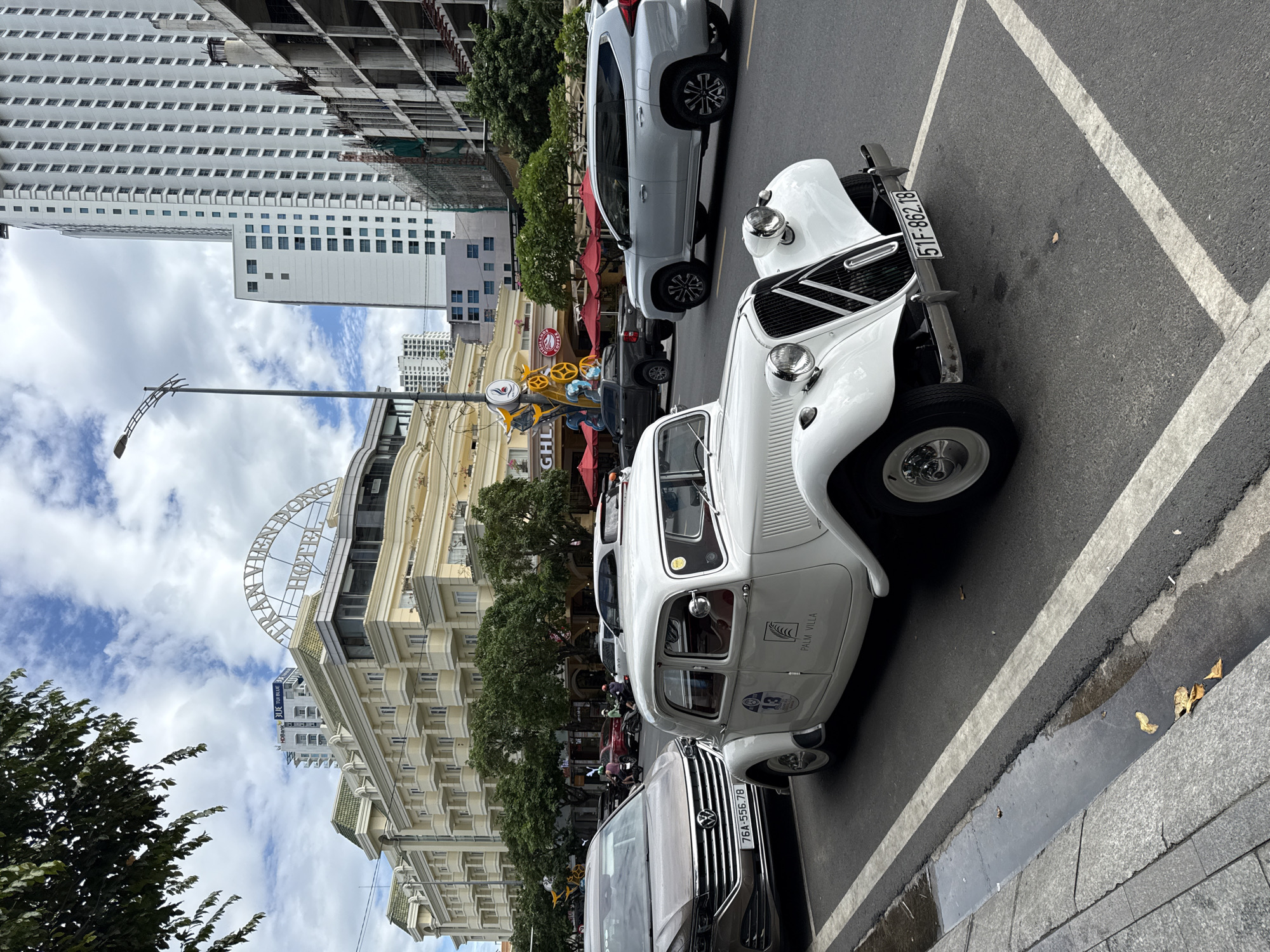 The image captures a street scene in Nha Trang, Vietnam, likely along Tran Phu Street, a prominent coastal boulevard. In the foreground, a vintage white Citroen car is parked at an angle. The car has a visible license plate reading "51F-862.18" and a circular emblem with the number "13" and "PALM VILLA" printed on the side. Beside it, a modern silver SUV is also parked.
Behind the vintage car, traffic is visible, including a brown Volkswagen SUV with the license plate "76A-556.78", a dark pickup truck, and several motorcycles with riders.
Dominating the background is the "TRAN VIEN DONG HOTEL," a large, light-yellow building with green accents and numerous balconies. Adjacent to it, to the right, are modern high-rise buildings, one of which appears to be under construction with exposed concrete framework. Red umbrellas are set up outside a commercial establishment with signs that are partially visible. The sky is cloudy, suggesting a typical day in the coastal city. The street is paved with asphalt, and a drainage gutter runs along the curb on the left side of the image, containing some puddles and fallen leaves.