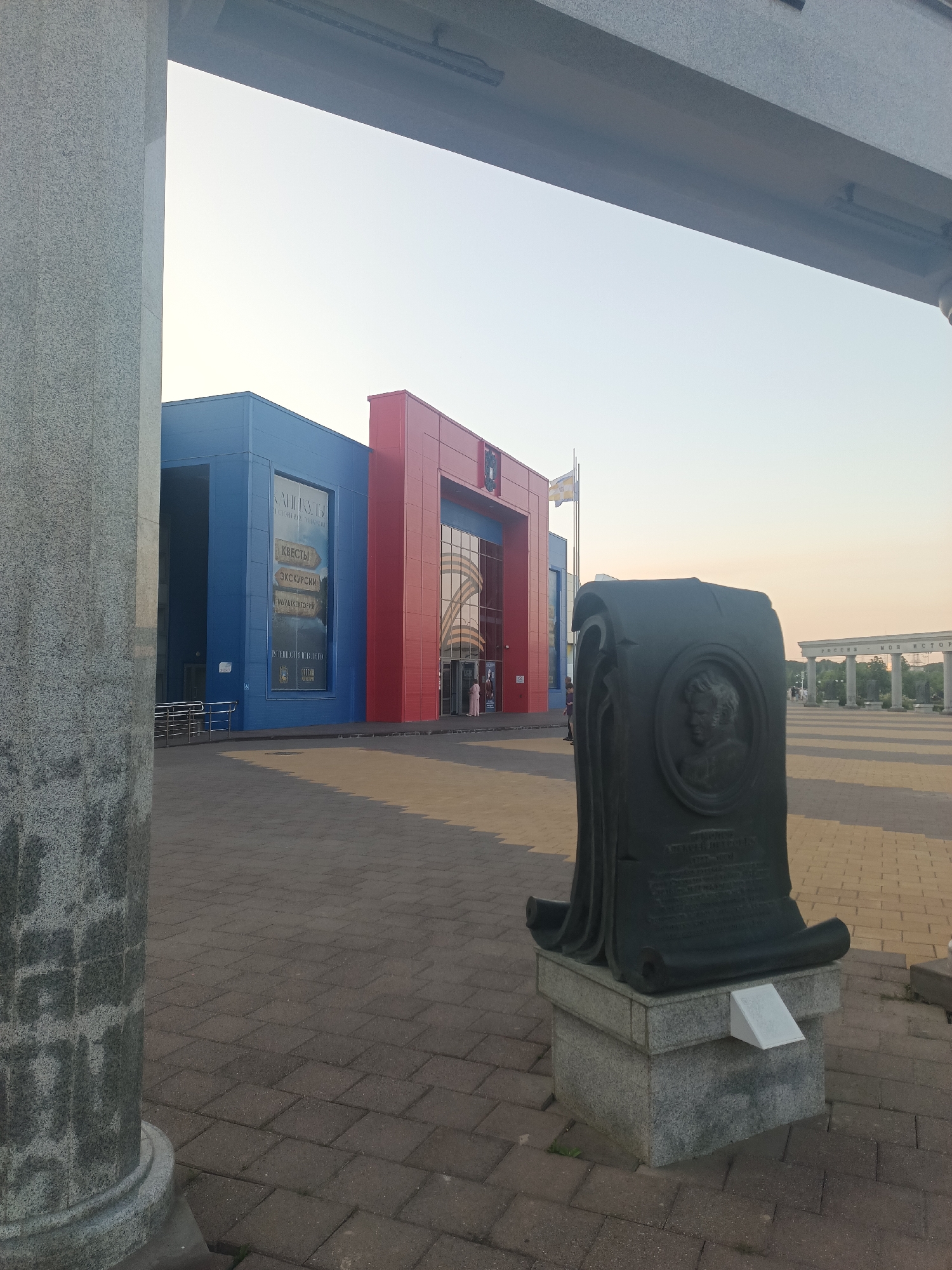 This image captures an outdoor scene in the city of Stavropol, Russia, likely within a public square or park. In the foreground, a dark, scroll-shaped monument with a circular relief portrait and an inscription stands on a stone pedestal. To the left, a large, grey, fluted column suggests an architectural element, possibly part of a building or colonnade.
Dominating the mid-ground is a modern, brightly colored building complex, featuring sections in vibrant blue and red. The blue section has large windows and displays signage, including text that appears to be in Russian. A flag is visible on a flagpole near the red section.
The ground is paved with interlocking rectangular stones. In the background, a series of classical-style white columns form an arcade or colonnade, with trees and a pale sky visible beyond. The overall lighting suggests it is daytime, with the sun low in the sky, casting shadows. A few indistinct figures can be seen in the distance near the building entrance and the colonnade, indicating a public space. The scene conveys a sense of civic monumentality and modern architecture.