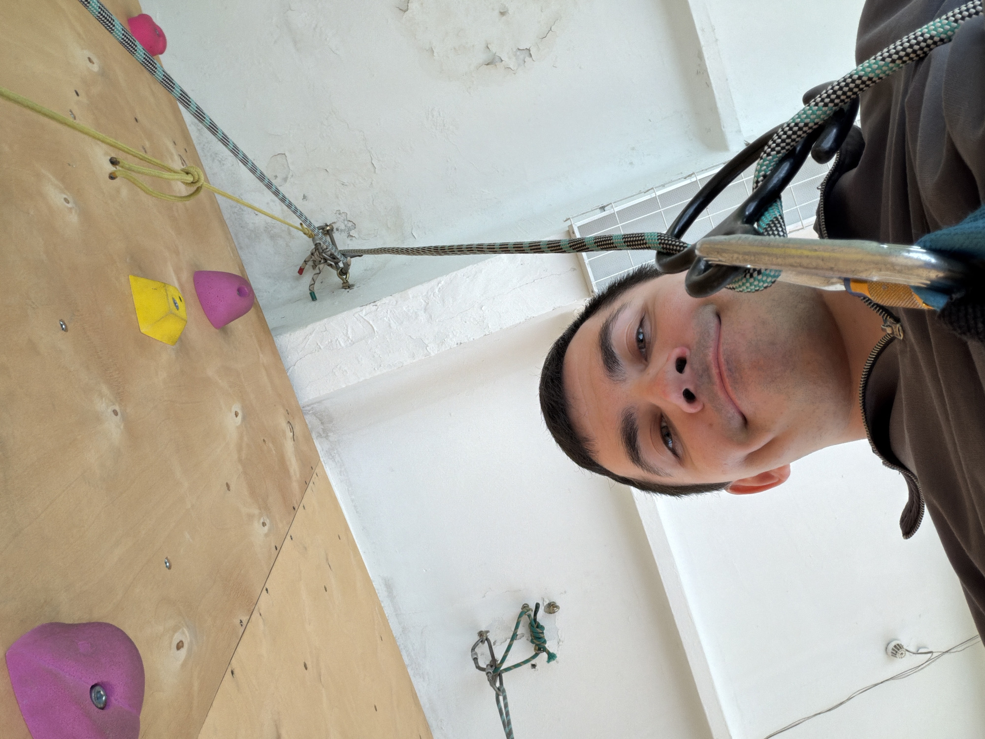 A man, seen from the nose up with a slight smile and direct gaze, is the central subject of this upward-angled close-up. He appears to be engaged in rock climbing, likely belaying or preparing for a climb, given the prominent climbing gear attached to him.
A multi-patterned climbing rope, featuring blue, black, and white, runs from the top of the frame, passing through a belay device and a large silver carabiner, which is attached to the man's person (though the harness itself is not visible). He is wearing dark gloves and a brown zip-up jacket.
To the left, a wooden climbing wall with various holds is partially visible. A bright yellow cubic hold and a vibrant pink conical hold are distinct on the wall. A yellow static cord or sling is knotted and connected to an anchor point on the wall or ceiling, leading towards other climbing hardware.
The background primarily consists of a white ceiling with noticeable peeling paint and some cracks, suggesting an older indoor facility. A metal grate or vent is also visible on the ceiling. Another anchor point with ropes is visible further along the ceiling.
The image captures an indoor rock climbing environment in Izhevsk, Russia, with a focus on the climber's perspective or interaction with the camera during an activity.
