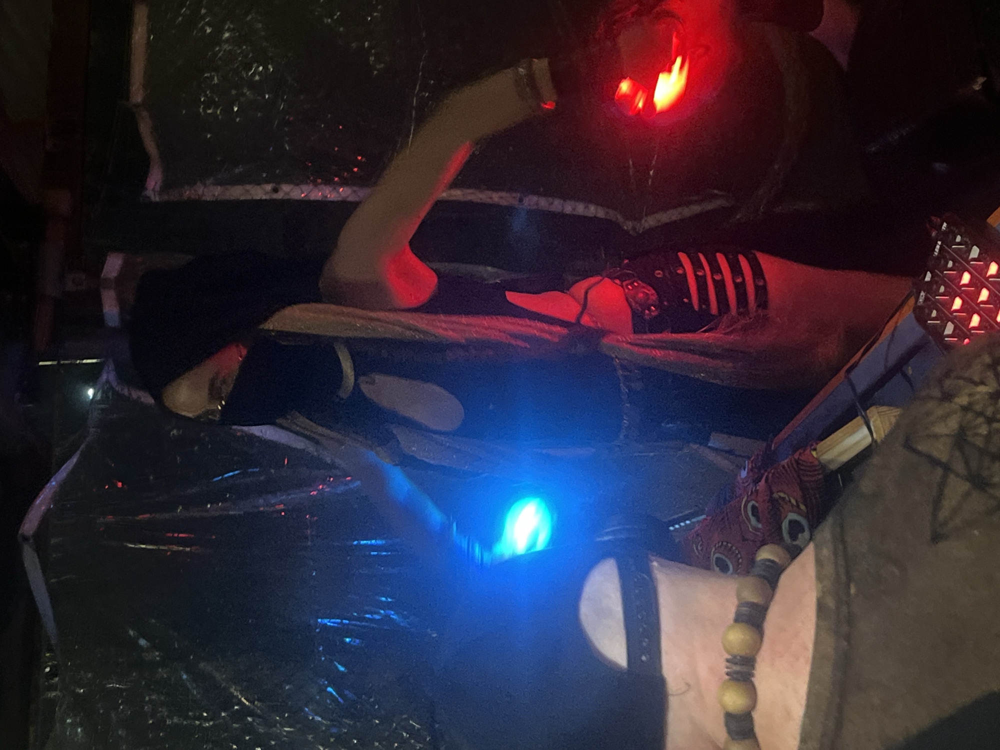 In a dimly lit indoor environment, possibly a haunted house attraction, a person wearing a black hood and a face mask is visible. They are holding two illuminated objects, one emitting a bright blue light in their left hand and another emitting a red light in their right hand. The person is dressed in dark clothing with cutouts on the chest and what appears to be a studded belt or accessory around their waist. The background is dark and appears to be decorated with reflective or shiny materials. There are other objects in the foreground and background, including what looks like part of another person's shoulder with a beaded necklace, and red lights illuminating parts of the scene. The overall atmosphere is eerie and theatrical. The image was taken in Đà Nẵng, Vietnam.