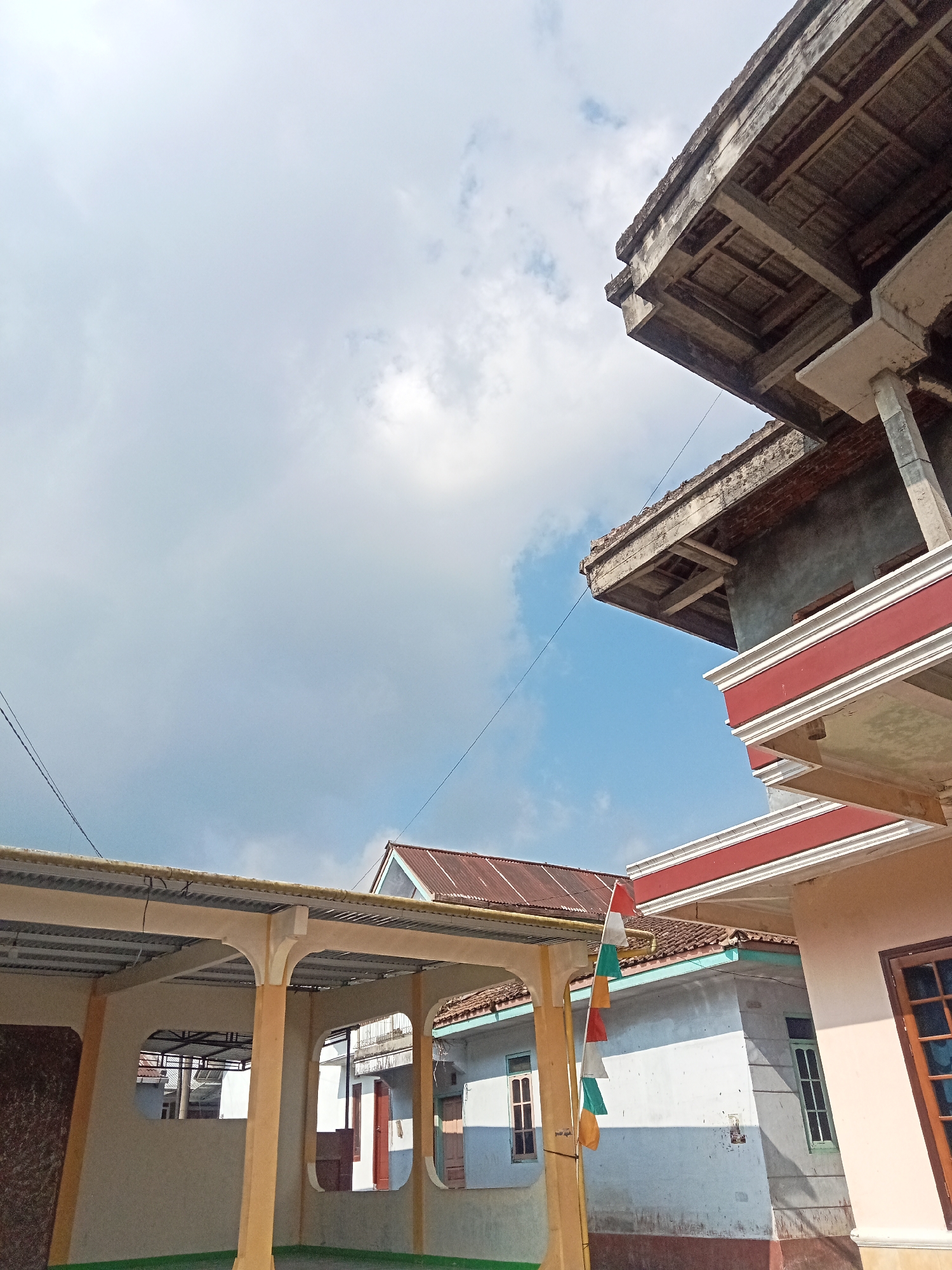 The image depicts a residential area under a bright, partly cloudy sky, likely in Pancurwening, Indonesia. The foreground features a covered outdoor space with yellow pillars supporting a corrugated metal roof. This area has open arched sections revealing other buildings and structures behind it. To the right, a two-story house is partially visible, characterized by its light pink exterior, prominent red and white decorative trim along the upper level, and wooden-framed windows. The upper part of this building appears unfinished or under construction, with exposed concrete and brickwork. Another building with a reddish-brown tiled roof and light blue siding is situated behind and to the left of the pink house. A string of small, colorful flags—red, white, green, and yellow—hangs from a pole in front of this blue and white building. The background is dominated by the sky with large, white cumulus clouds against a clear blue backdrop. Electrical wires are visible crisscrossing the sky, indicating an urban or suburban setting. The overall impression is one of everyday life in a community with both completed and ongoing construction.