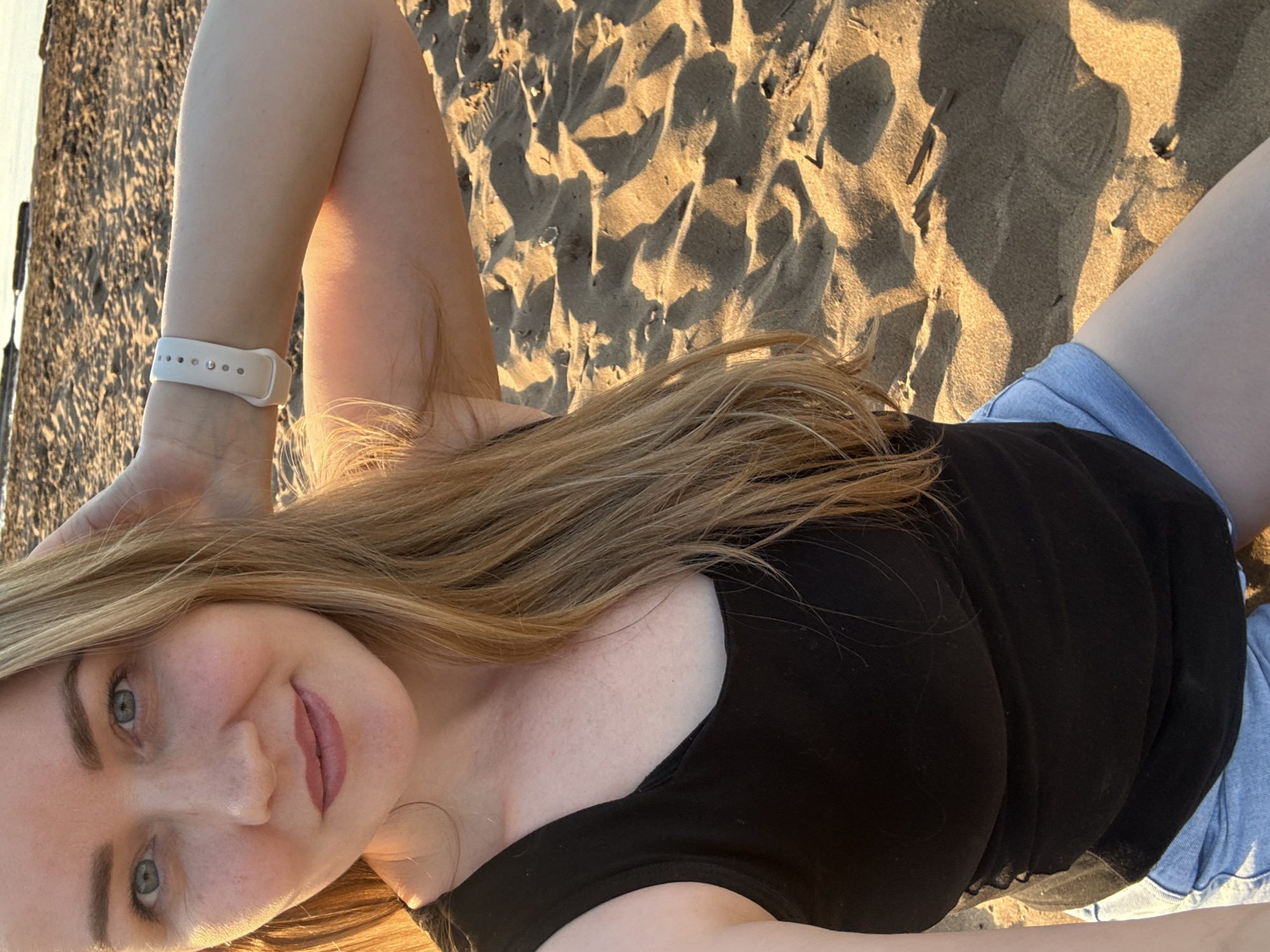 The image shows a woman lying on a sandy beach. She has blonde hair, light skin, and is wearing a black tank top and denim shorts. A white smartwatch is visible on her left wrist. The sand is light brown, and there are shadows cast across it, suggesting a sunny day. In the background, a blurred structure is visible along a body of water. The location is Chaikovsky, Russia.