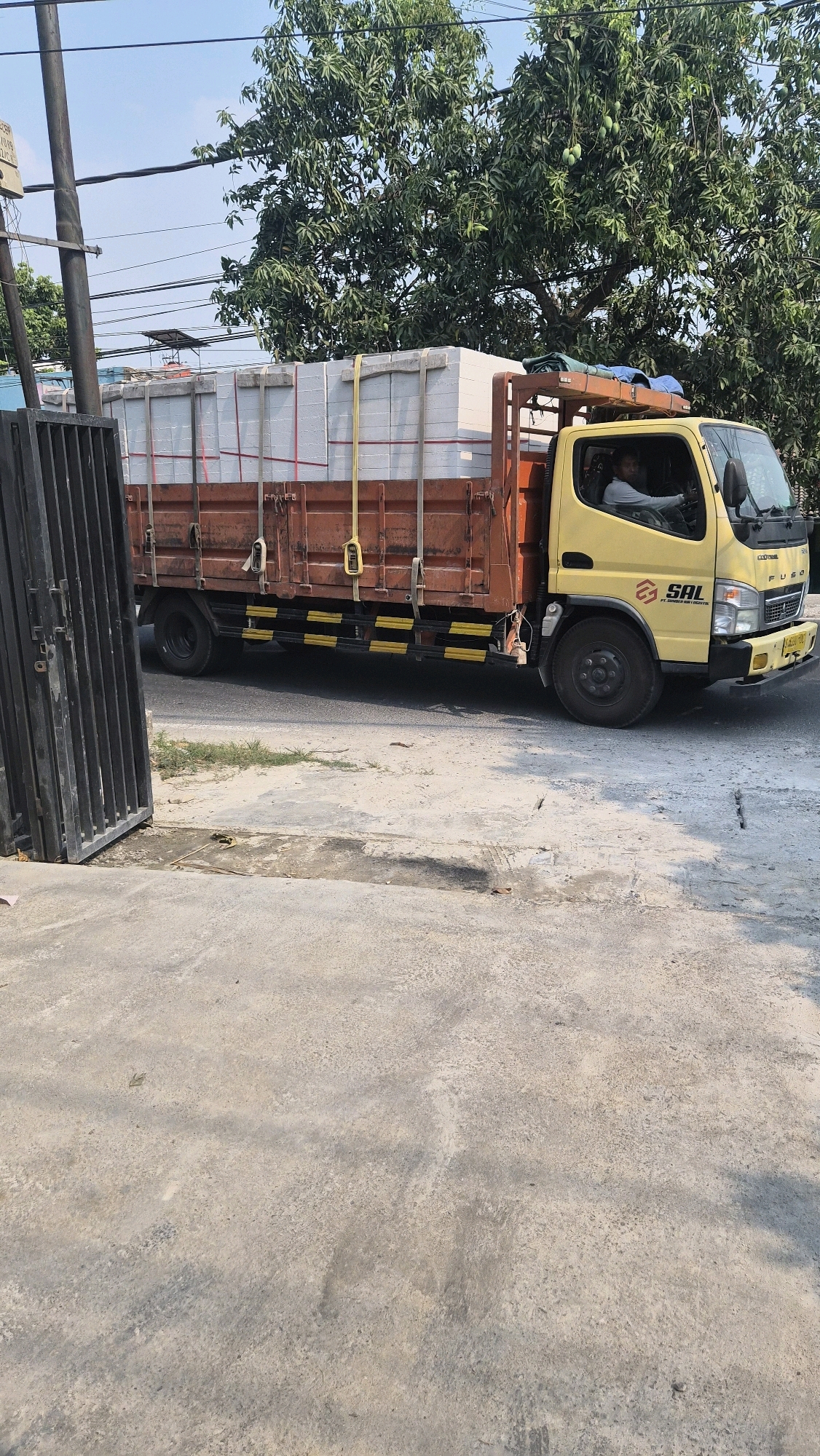 A yellow Fuso truck, laden with white rectangular objects secured by straps, is parked on a street. The truck has "SAL" logo and "FUSO" branding visible. A man wearing a light-colored shirt is seated in the driver's seat. The truck is next to a dark metal gate. The background features a large tree with green foliage and power lines overhead, suggesting an outdoor, possibly semi-urban or industrial setting. The ground is paved with concrete. The location is identified as Mernung, Indonesia.