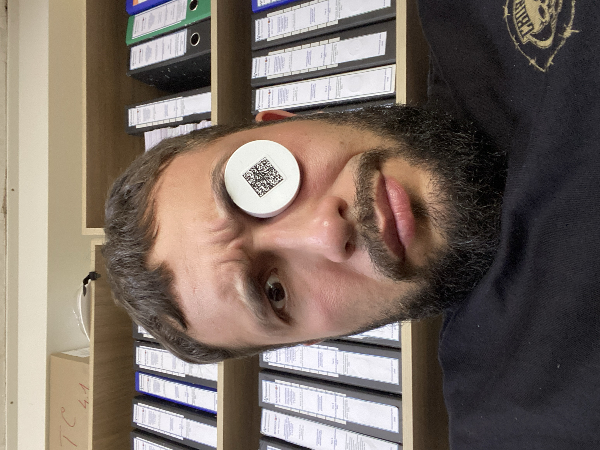 The image shows a close-up selfie of a man with a beard. He is wearing a black t-shirt and has a white circular object with a QR code on it placed over his left eye. He is looking directly at the camera with a somewhat surprised expression. Behind him, there are shelves filled with binders. The man is positioned in an office environment. The location is identified as Luzhitsy, Russia, suggesting an interior setting within a building in that city.