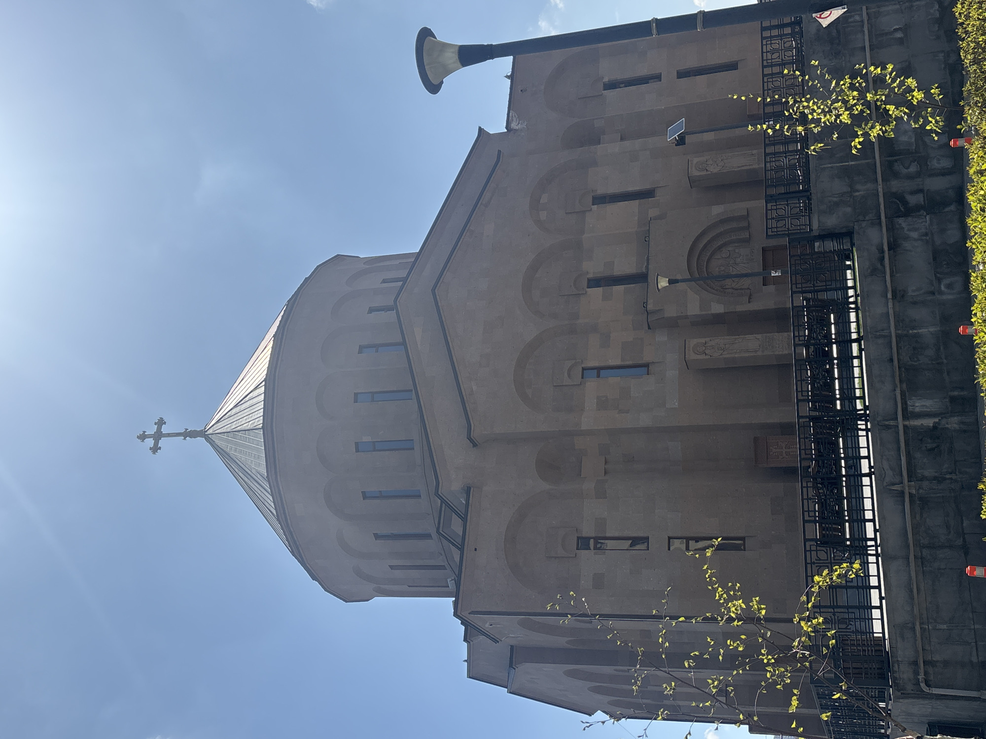 The image depicts the Saint Gregory the Illuminator Cathedral in Yerevan, Armenia. The cathedral is constructed of a light-colored stone. The architecture includes a central dome topped with a cross and several arched features. A black metal fence is located to the right of the cathedral. A tall, black pole with a speaker-like device attached to the top is also visible. A young tree with green leaves is present in the foreground. The sky is blue with a few clouds and sunlight.