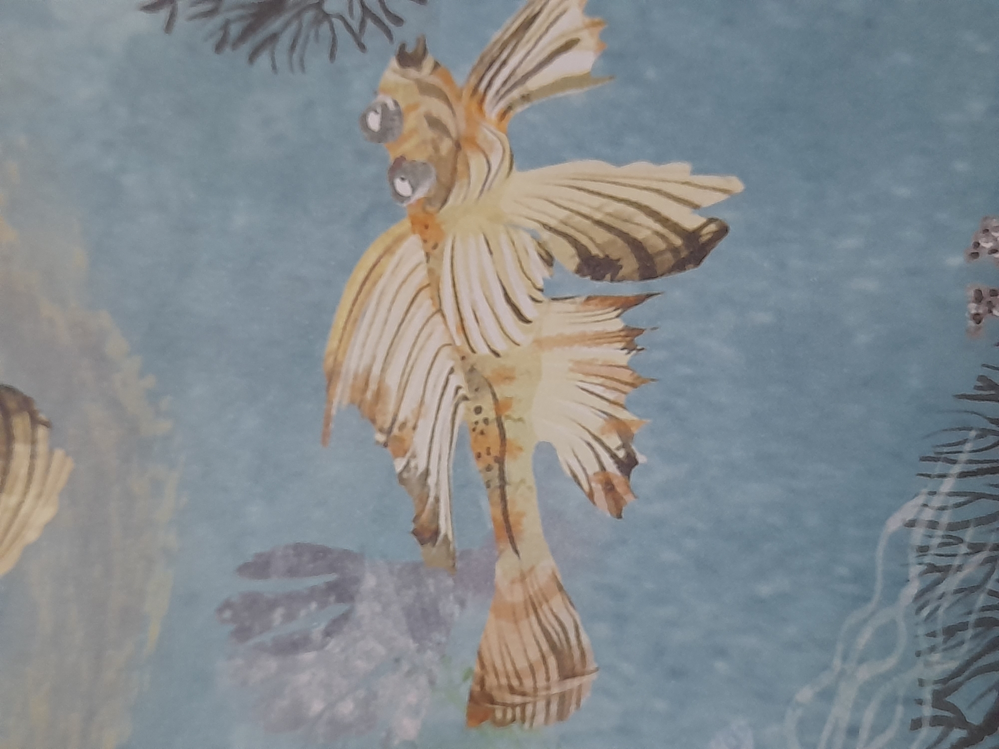 The image is a close-up shot of a decorative pattern on a blue background. The pattern features a stylized depiction of a yellow and brown fish with large, bulging eyes. The fish has prominent fins and tail, with dark stripes accentuating its form. The background appears to be a textured wallpaper or fabric, with hints of other abstract shapes and elements that might represent underwater flora or other marine life. There are no people or identifiable landmarks from Baan Mae Nam, Thailand, in this image.