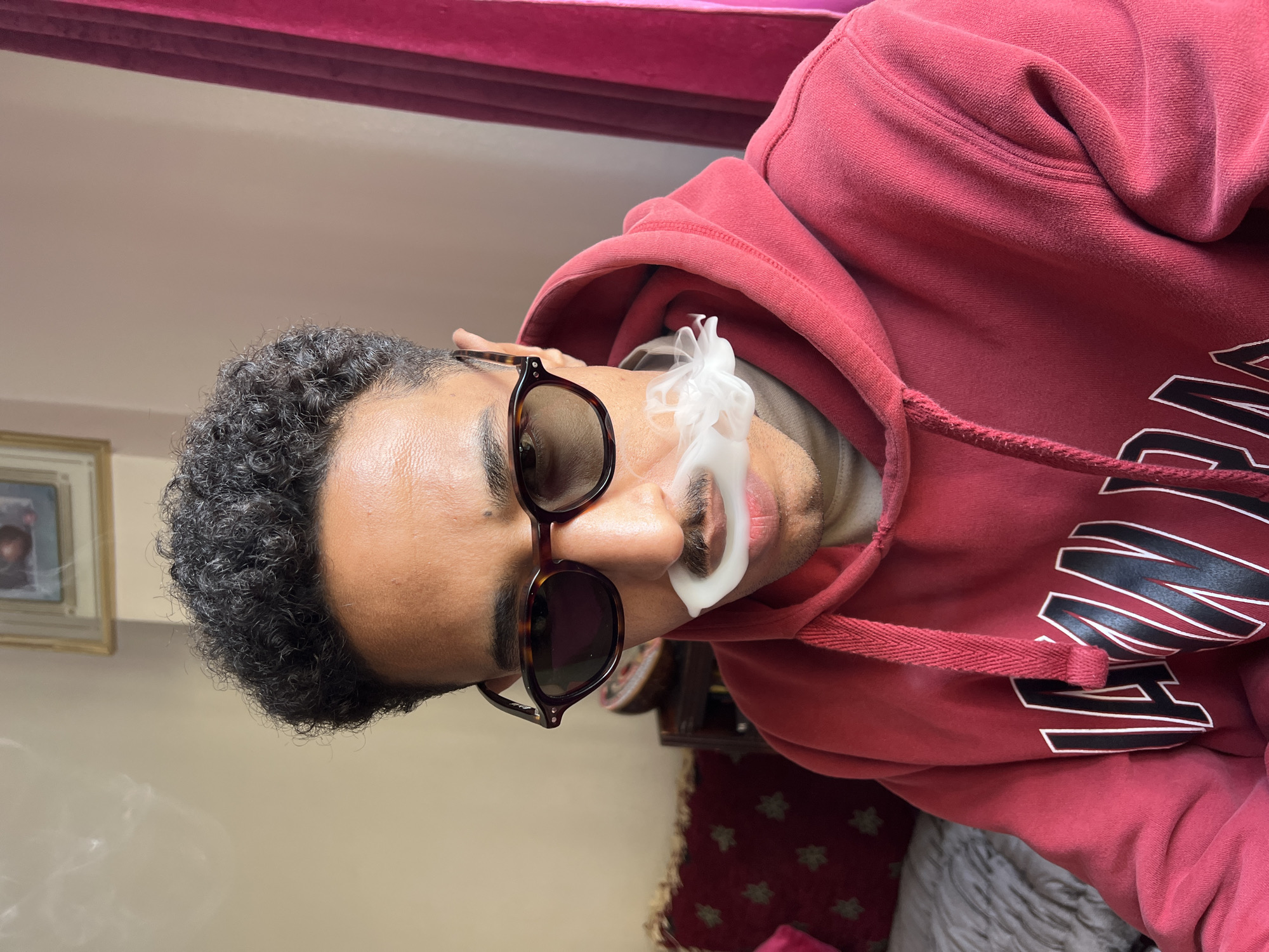 A man with dark curly hair and brown skin is wearing tortoise-shell framed sunglasses and a red hooded sweatshirt. He is exhaling a plume of white smoke from his mouth. The setting appears to be indoors, with a red velvet headboard visible at the top and a patterned cushion partially visible at the bottom. There is no evidence to determine if the location is Tanger, Morocco.